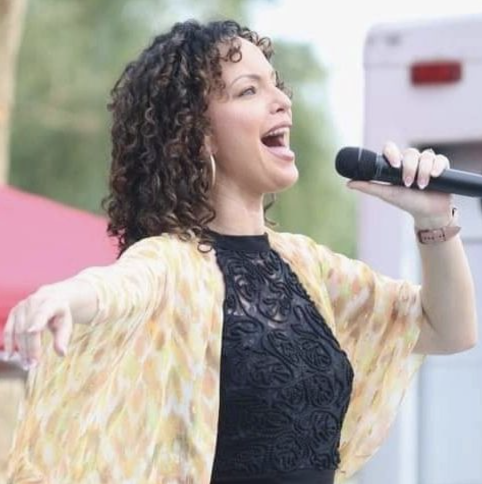 A woman singing into a microphone with her arms outstretched, wearing a black lace top and a sheer yellowish cover-up, outdoors with blurred background.