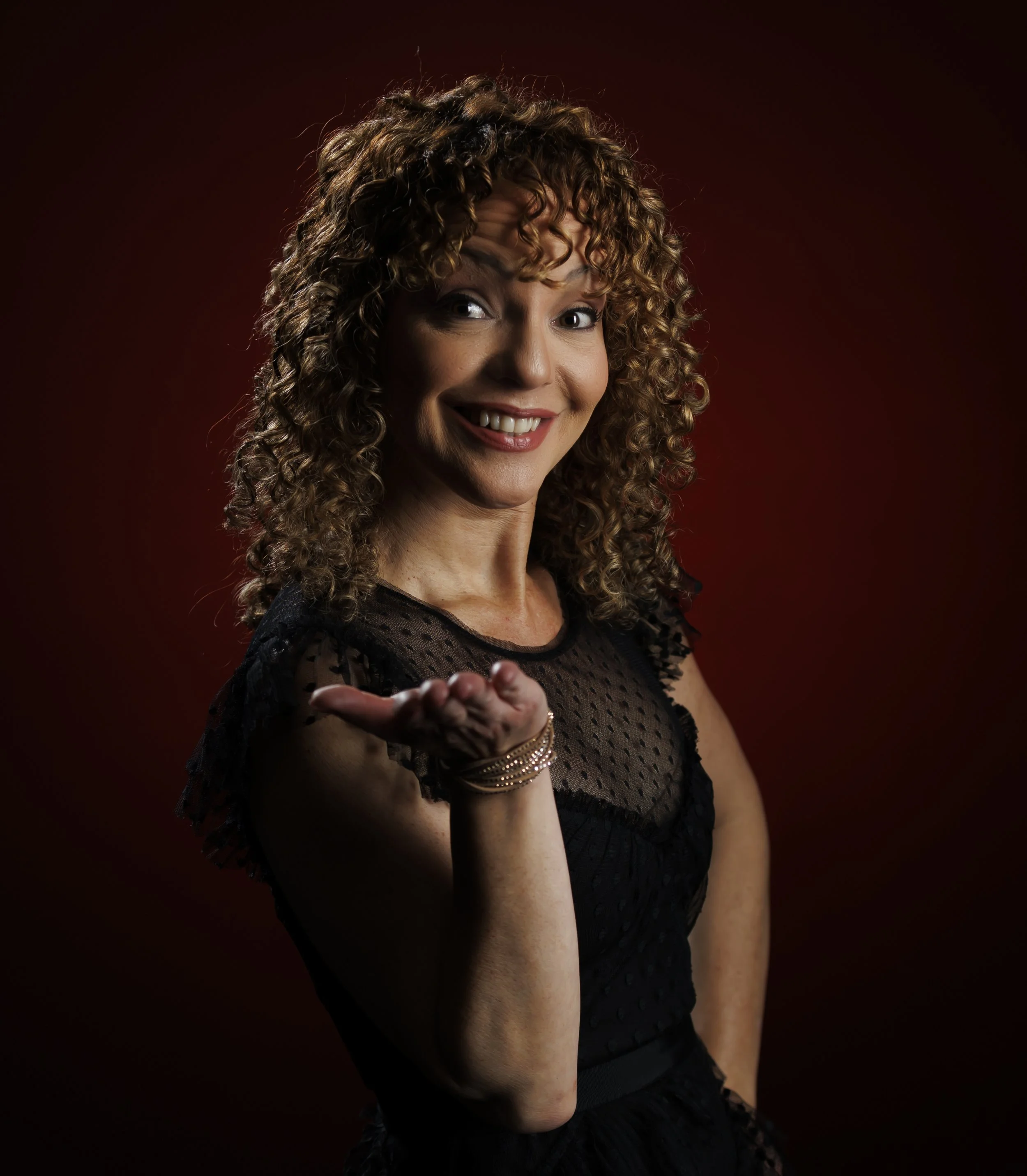A woman with curly hair, wearing a black dress with a dotted pattern, blowing a kiss toward the camera with a smile, against a dark red background.