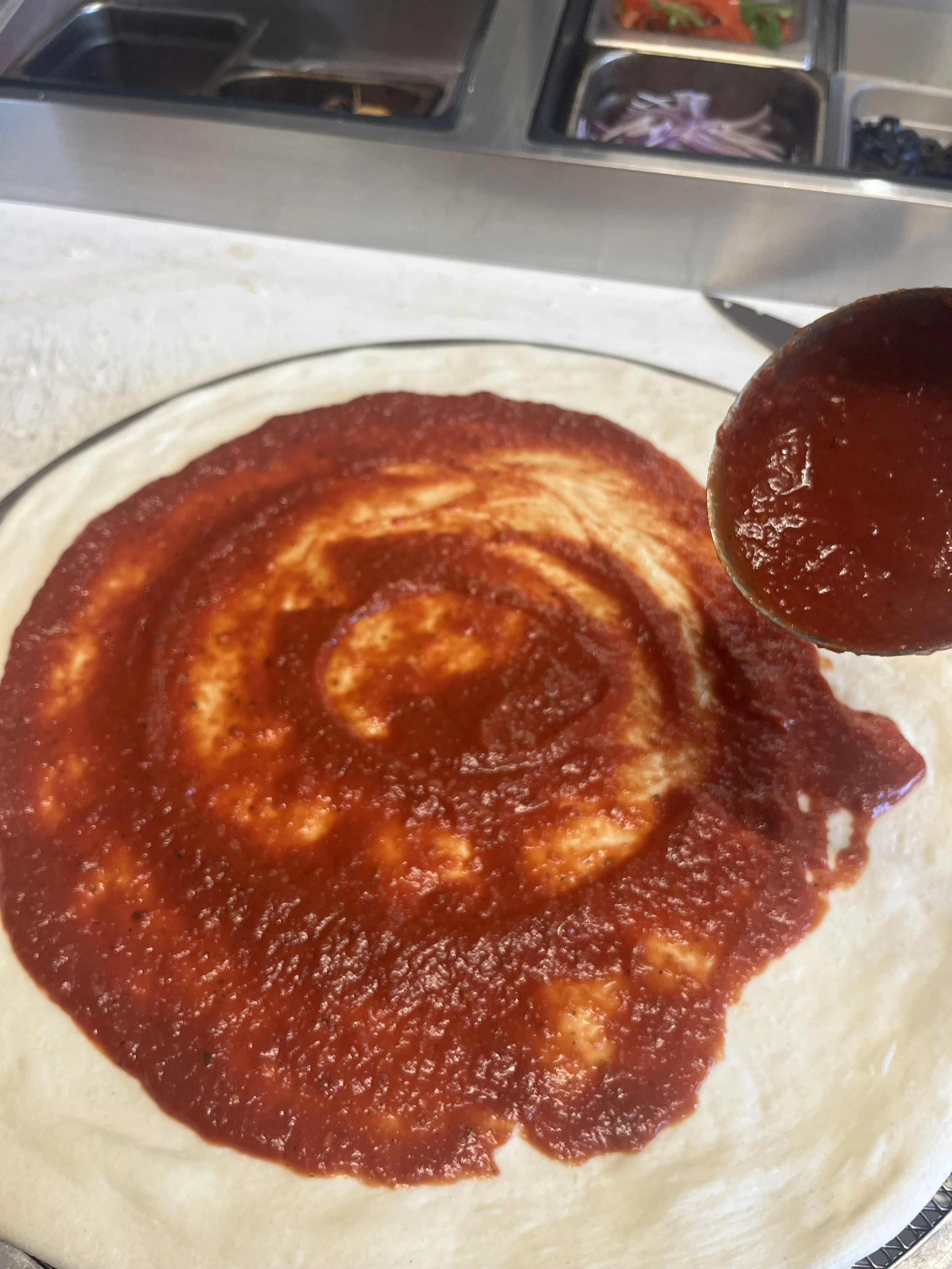 A hand-stretch sourdough crust with organic, housemade tomato sauce, almost ready to be topped. 