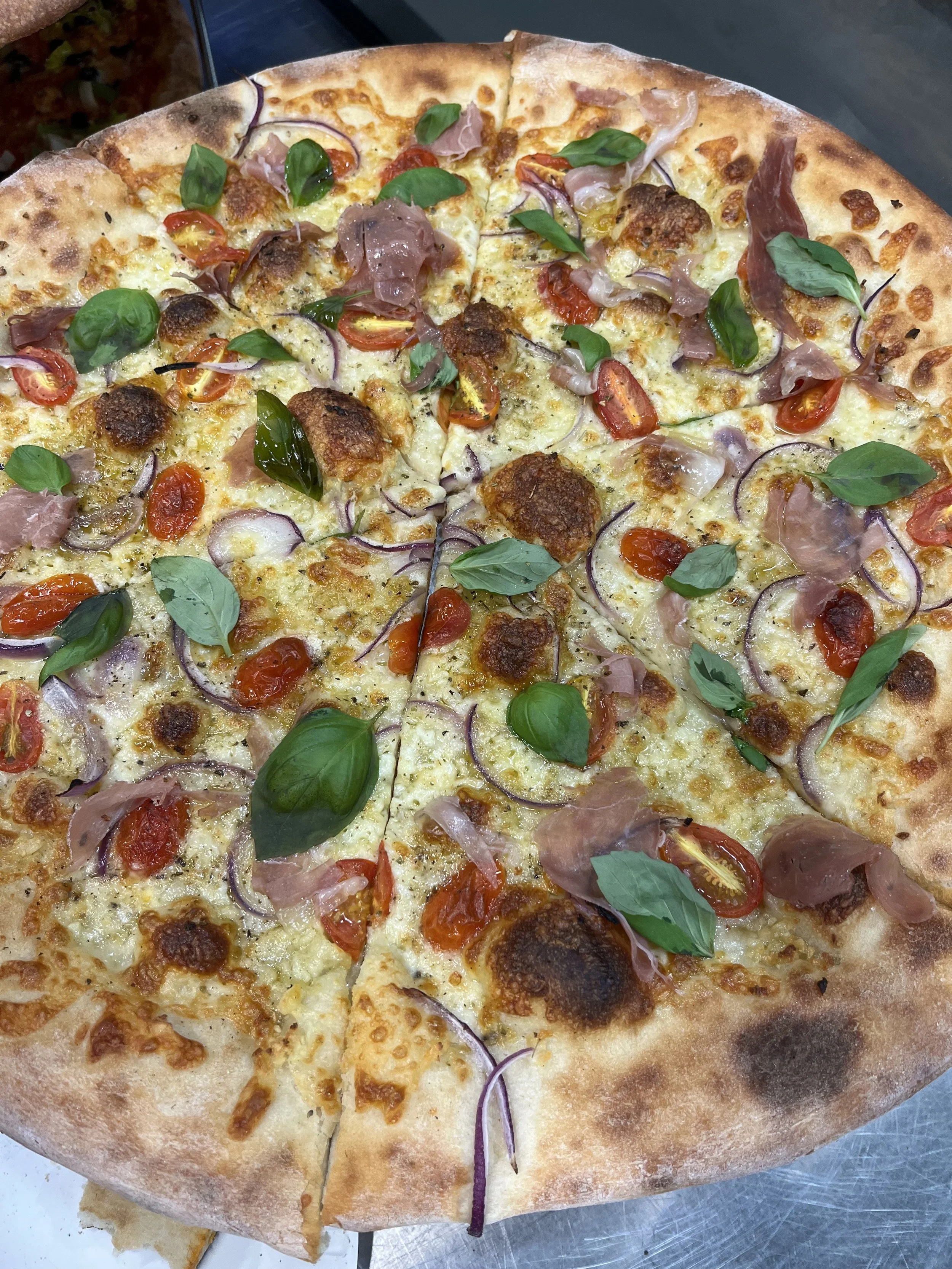 A freshly baked specialty pizza topped with cherry tomatoes, red onions, basil leaves, and slices of prosciutto, with a golden crust.