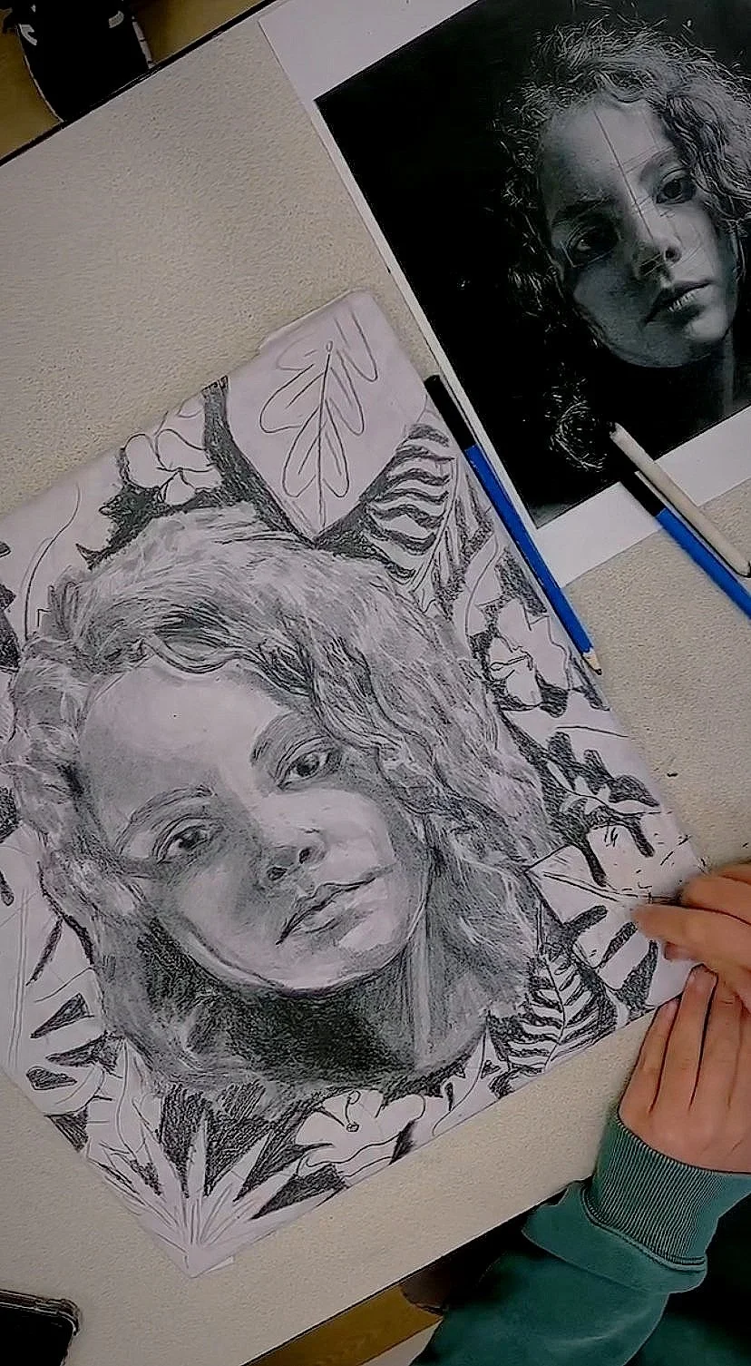 A pencil drawing of a young woman with wavy hair on a table, next to a reference photo. The drawing includes floral and leaf patterns in the background. Hands visible on the table.