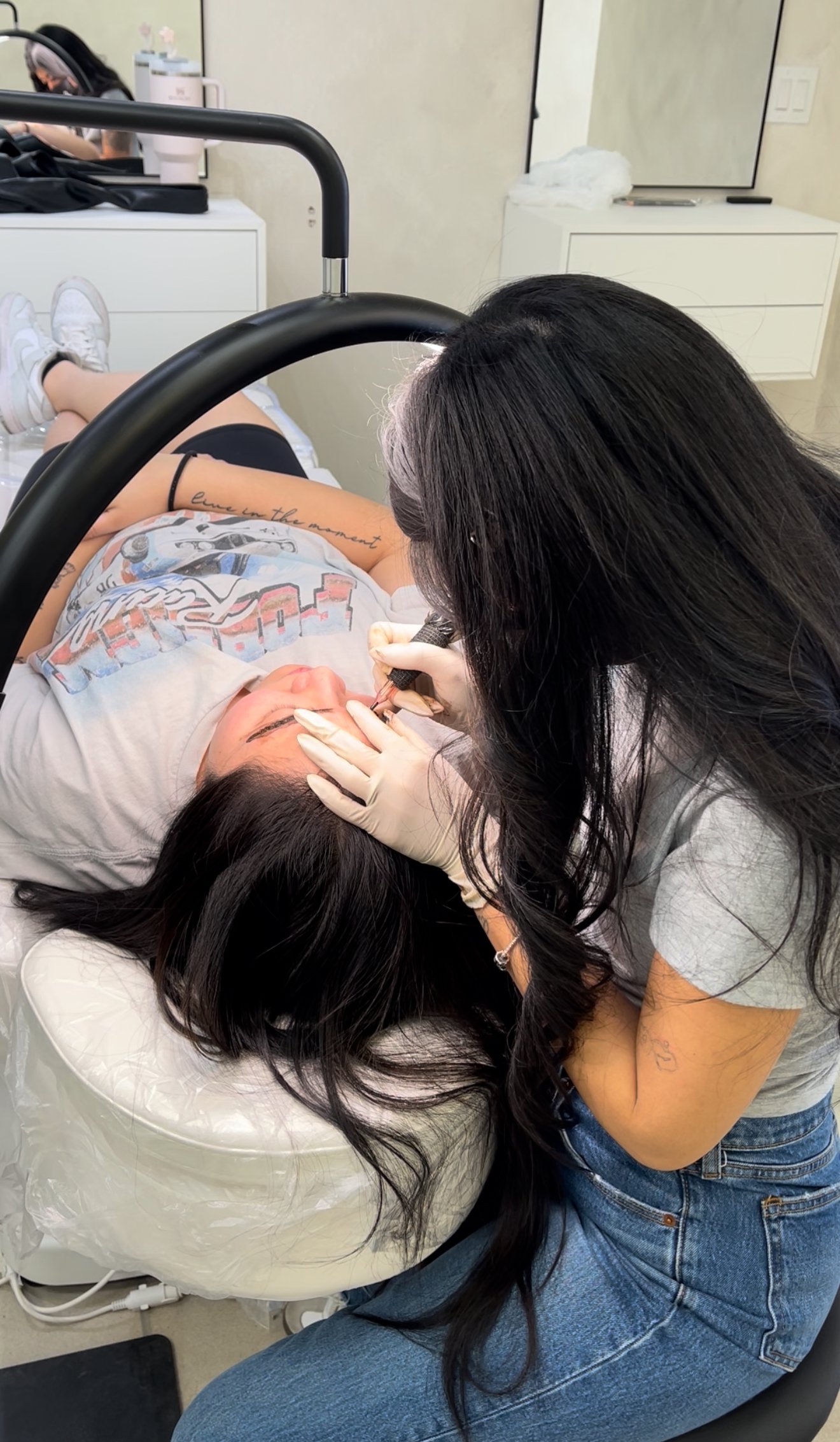 A cosmetologist performing a microblading procedure on a client's eyebrows, with the client lying down on a chair. The cosmetologist is wearing gloves and holding a microblading tool. The setting is a modern salon. Nano brows, PMU, permanent makeup