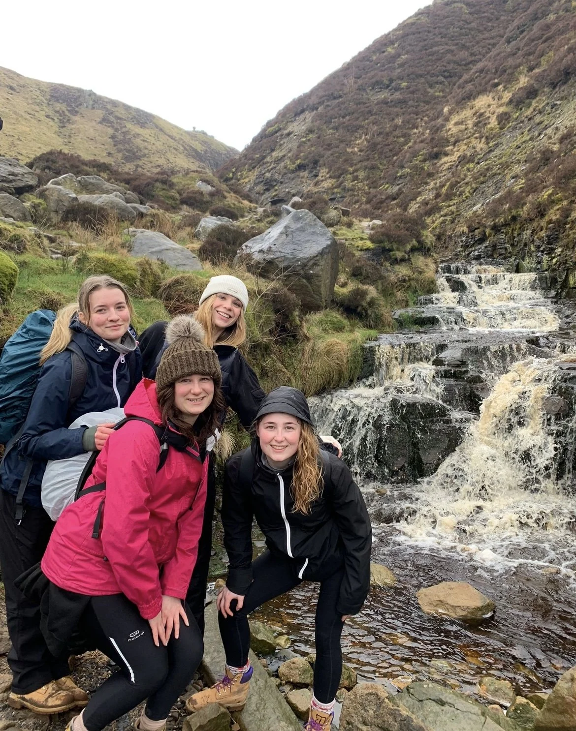 Four women hiking in a mountainous area near a waterfall, dressed in outdoor gear and smiling at the camera.