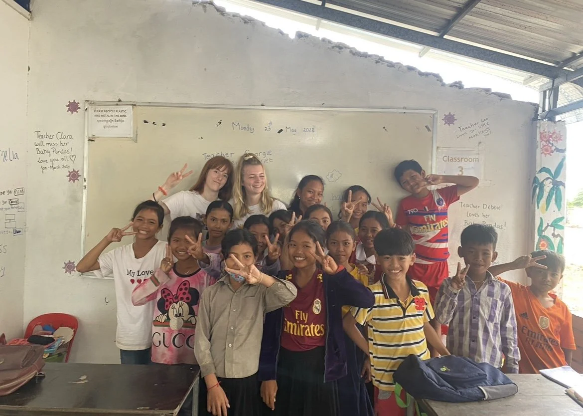 Group of smiling children and teachers in a classroom, some making peace signs, with a whiteboard and classroom wall in the background.