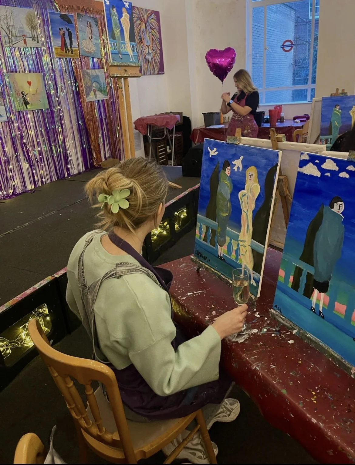 A woman sitting at a table in an art studio, holding a glass of champagne, observing two paintings of women on easels. The studio has colorful paintings, purple and gold tinsel decorations, and a pink heart-shaped balloon. A blonde woman with a green