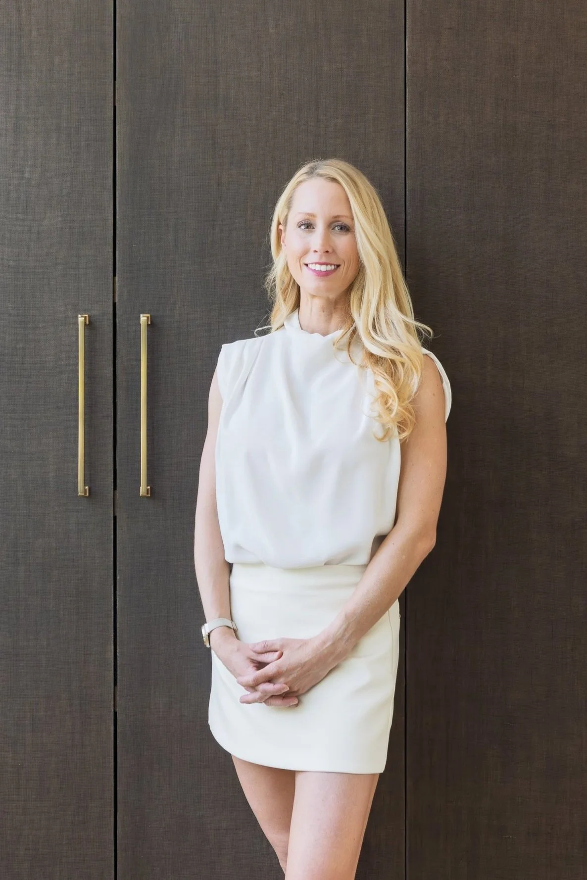 A woman with long blonde hair wearing a white sleeveless blouse and a matching white skirt, standing in front of a dark brown wooden cabinet with gold handles, smiling and looking at the camera.