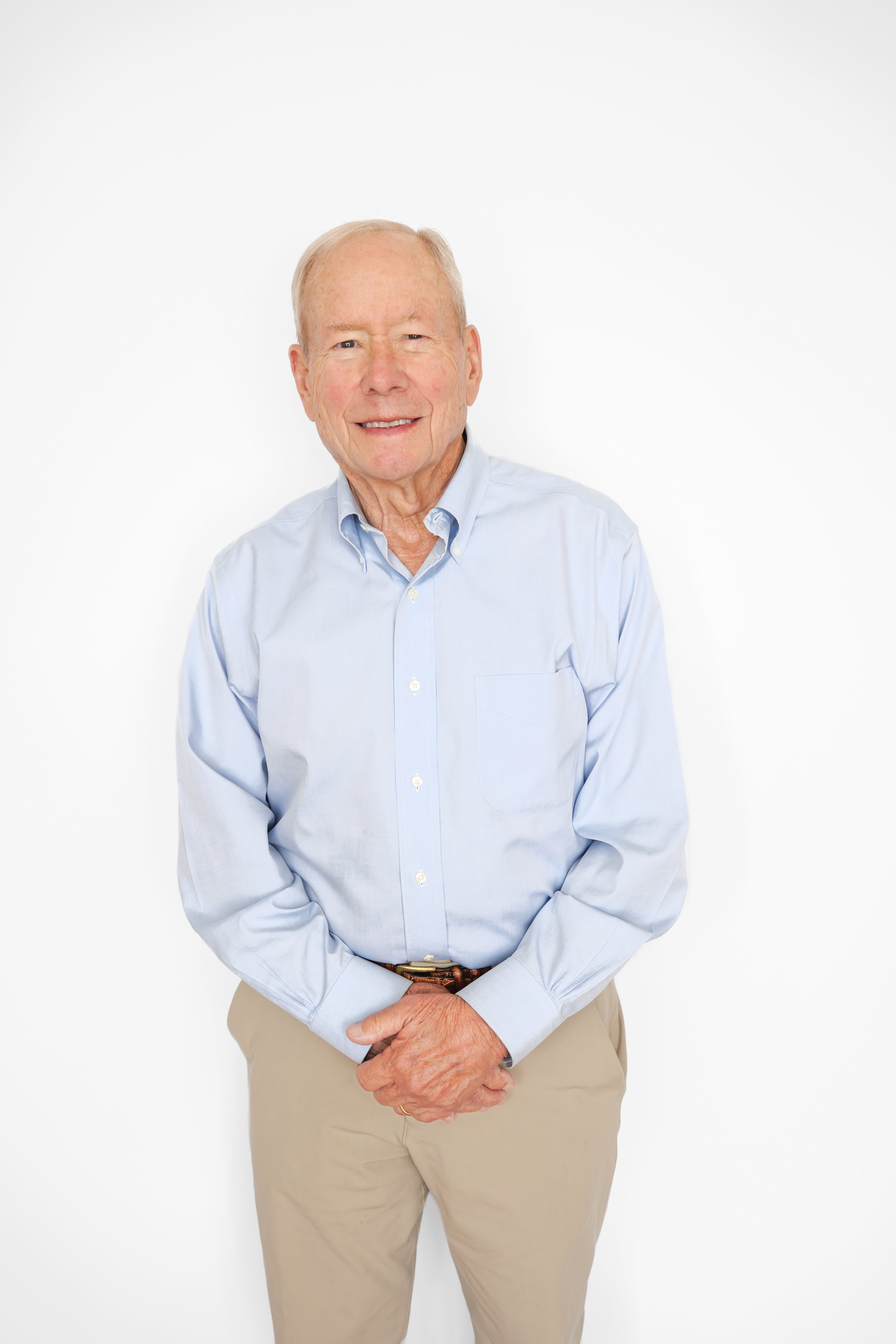 Older man with light skin, short white hair, wearing a light blue button-up shirt and khaki pants, smiling against a plain white background.