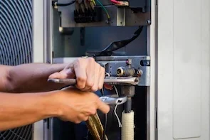 A plumber repairing or servicing the inside of an HVAC or electrical unit, using a wrench with various wires and components visible.