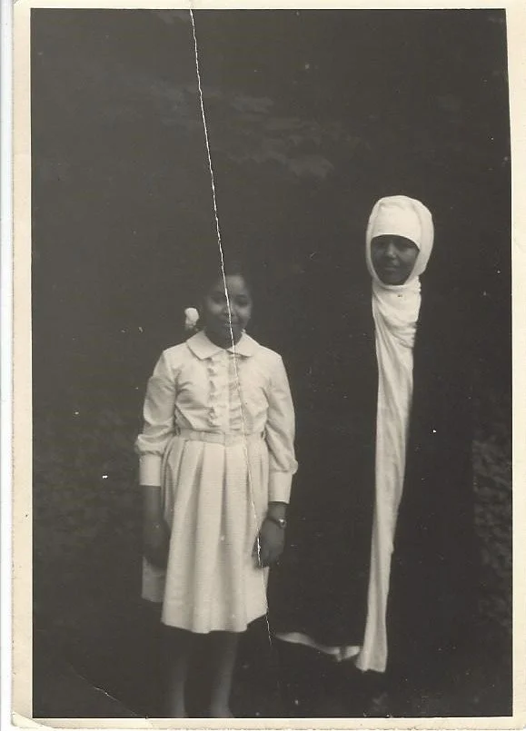 A young girl in a light-colored dress and a woman in a long black and white garment with a head wrap standing outdoors.