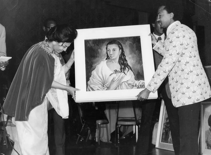 Three people holding a framed picture of a woman with long hair, dressed in white, at an indoor event.