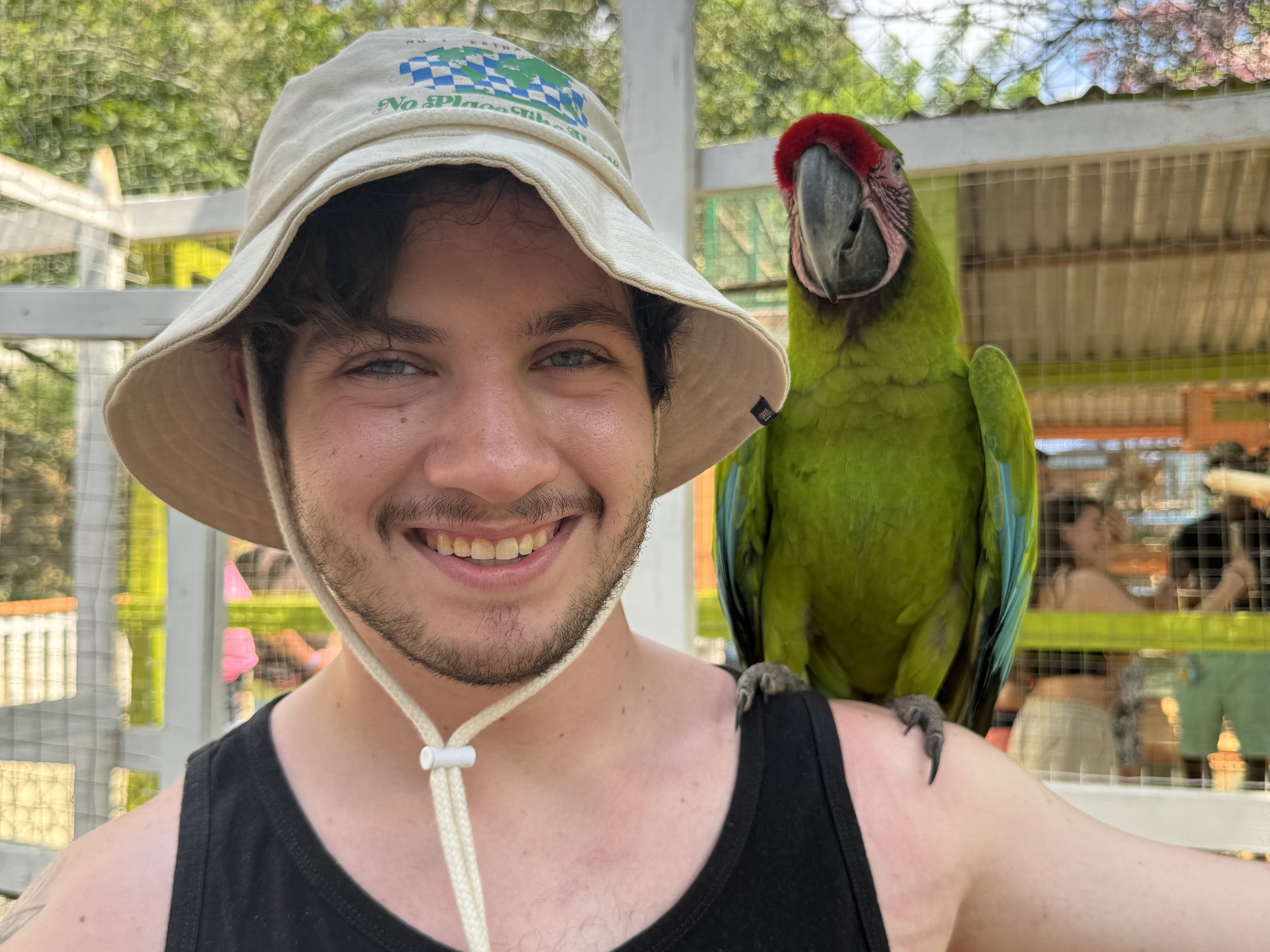 A smiling man wearing a sun hat with a parrot perched on his shoulder at an outdoor aviary or zoo.