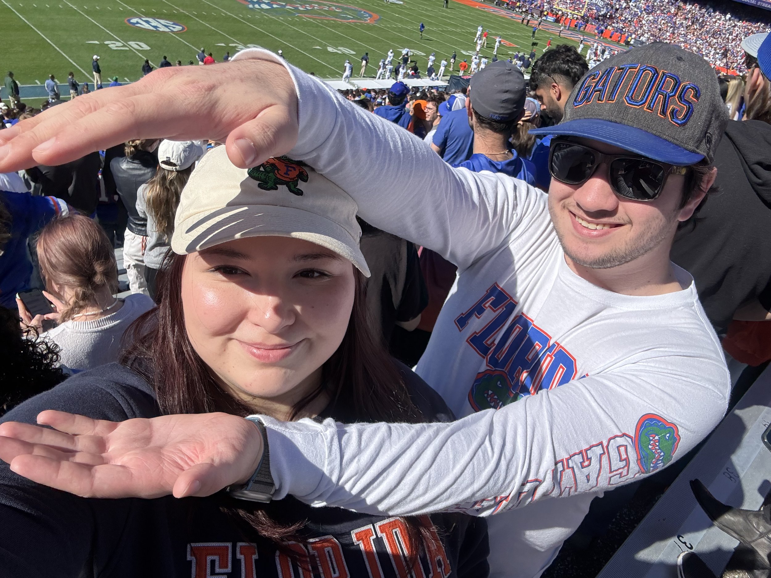 A man and woman at a football game, wearing Florida Gators clothing and hats, taking a selfie with the football field in the background.