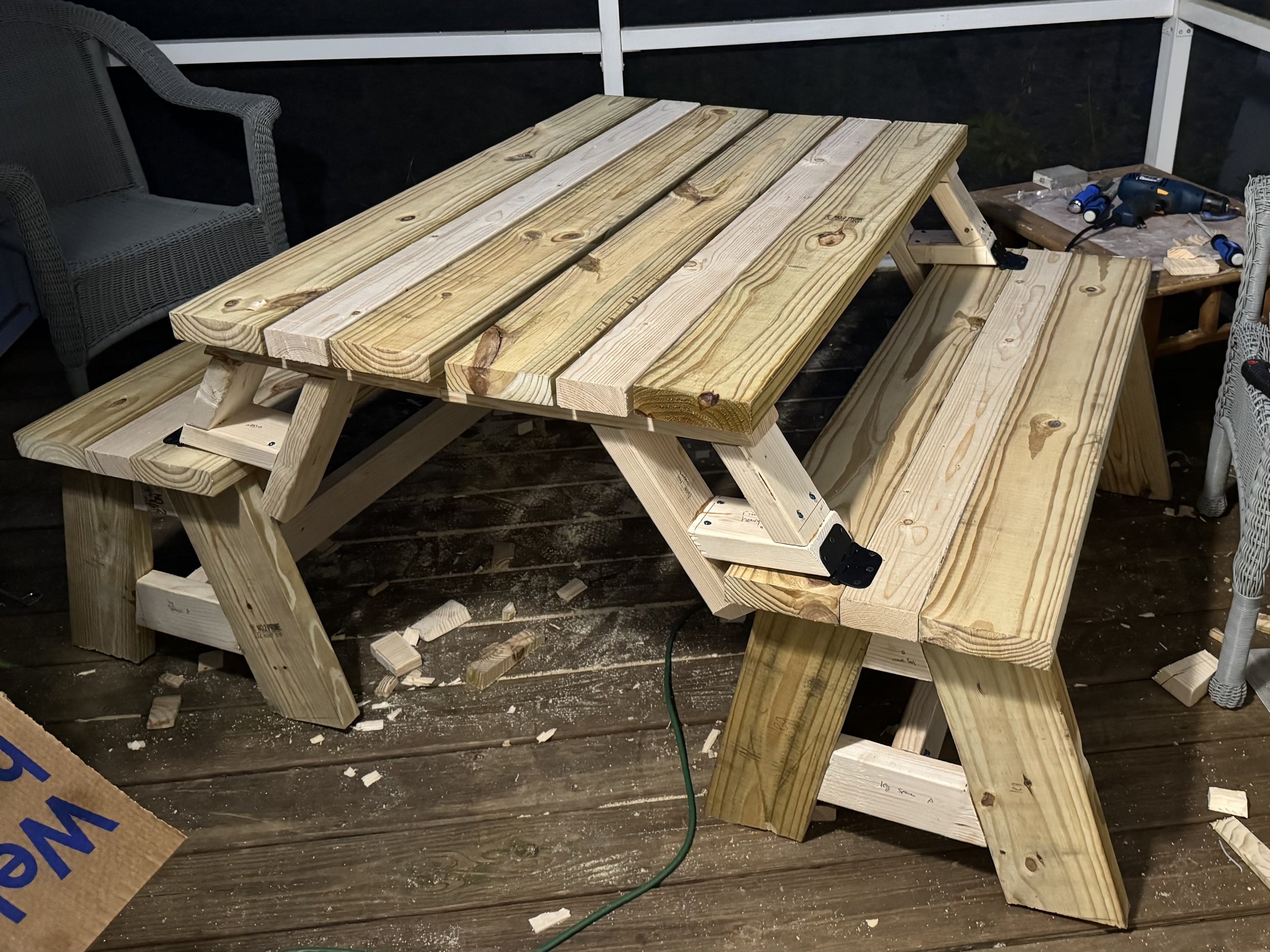 Unfinished wooden picnic table under construction on a wooden deck, with various tools nearby.