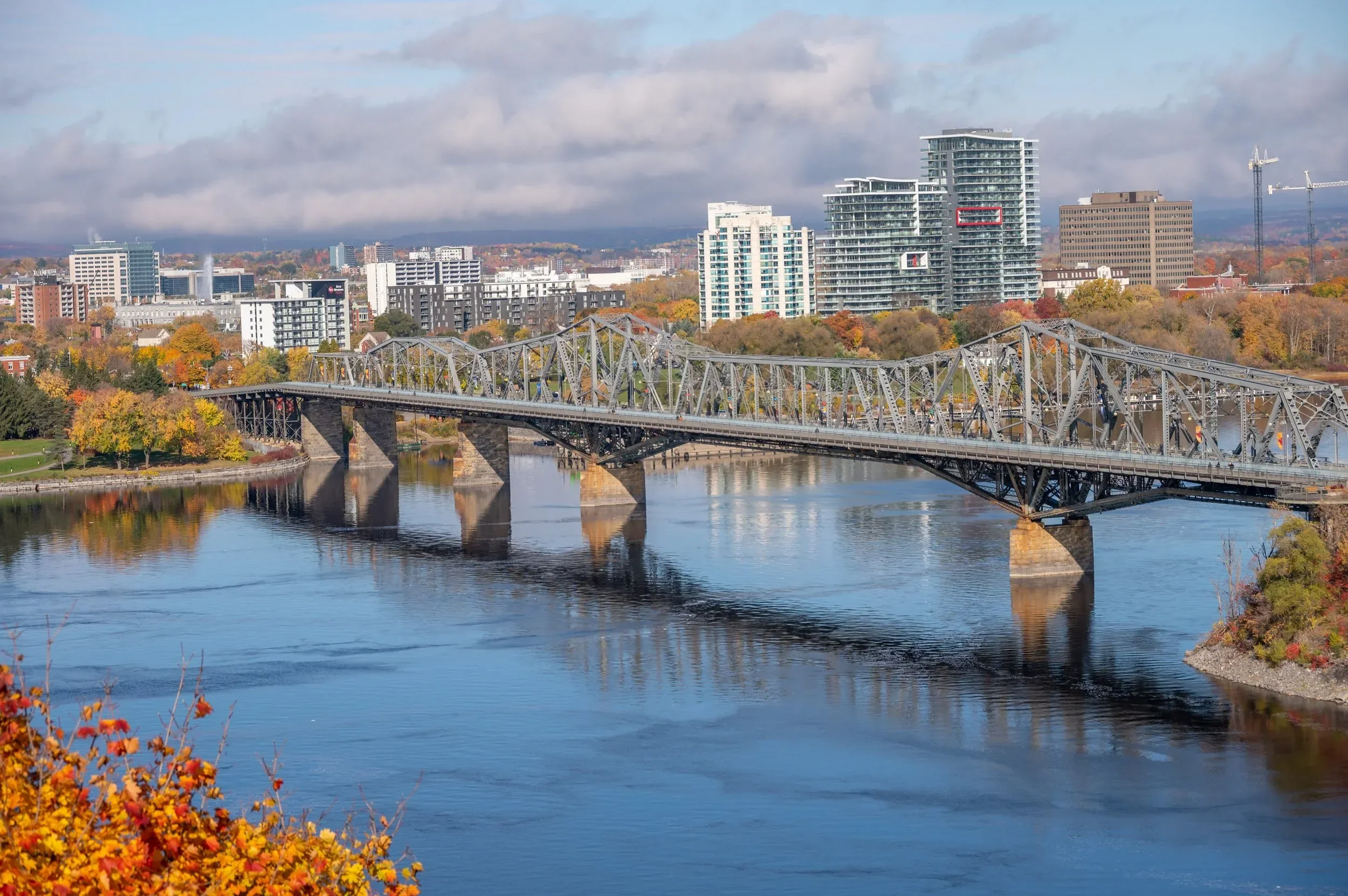A cityscape featuring a bridge over a river with modern high-rise buildings and trees with autumn foliage in the background.