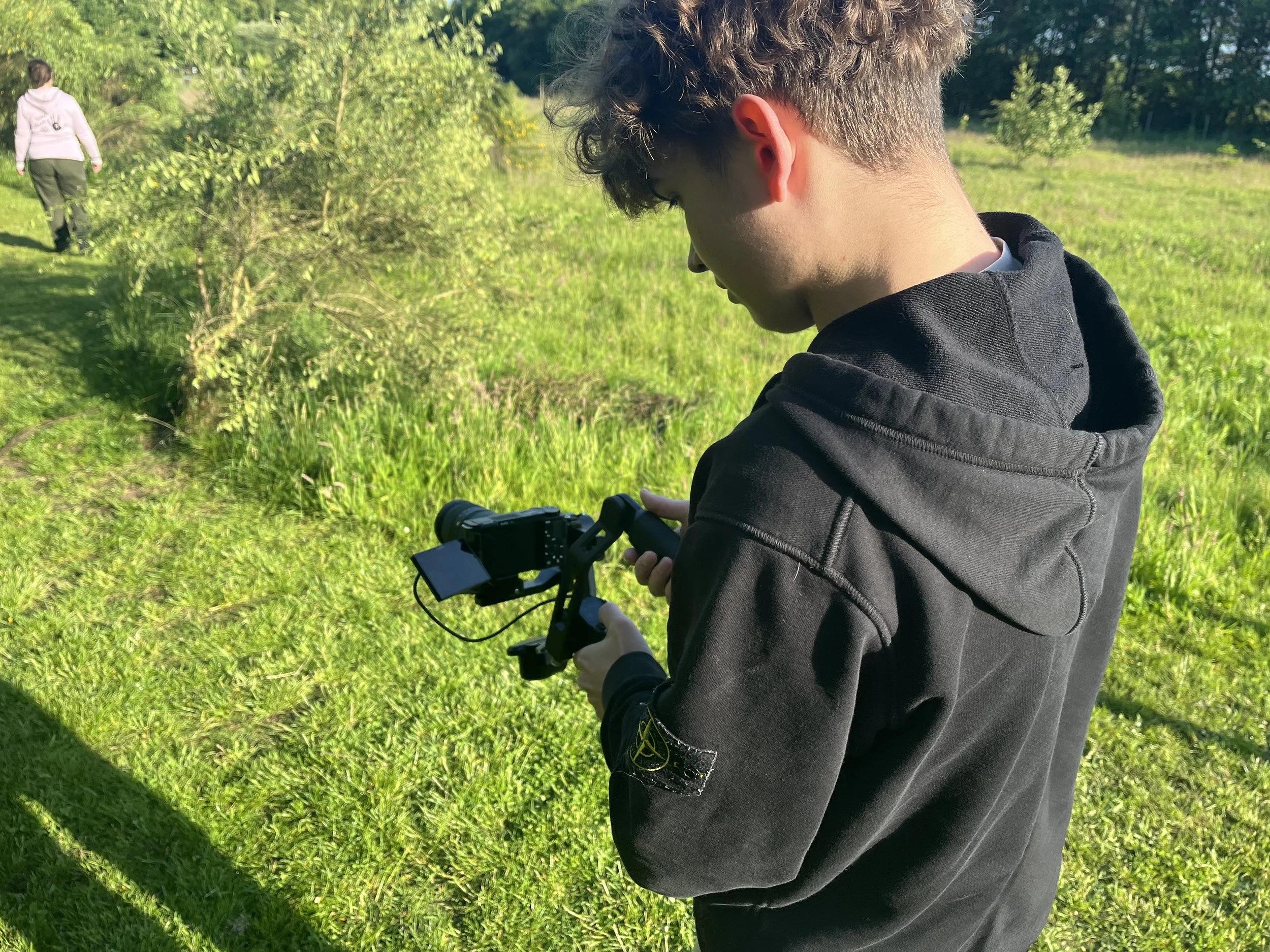 Ung mand i sort hoodie, der bruger en stabilisator, står i et grønt åbent område, og holder et kamera. En person går væk i baggrunden.