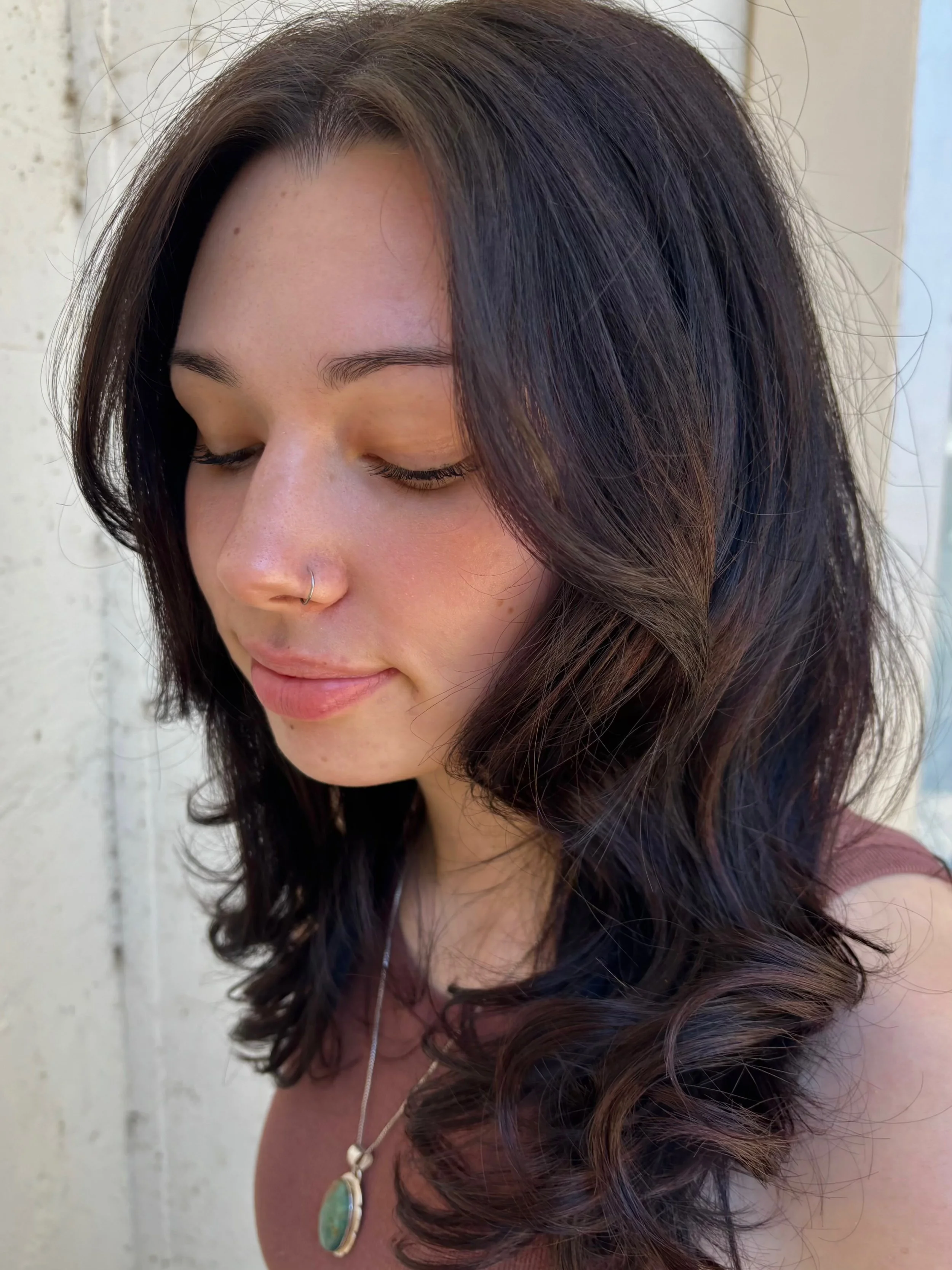 A young woman with long, dark, curly hair and fair skin, looking down with her eyes closed. She has a nose piercing and is wearing a necklace with a green pendant and a sleeveless top. She is standing outdoors near a white wall.