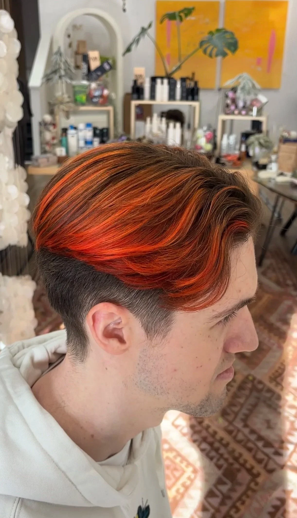 Young man with vibrant orange and red hair, styled in loose waves, at a hair salon with beauty products and artwork in the background.