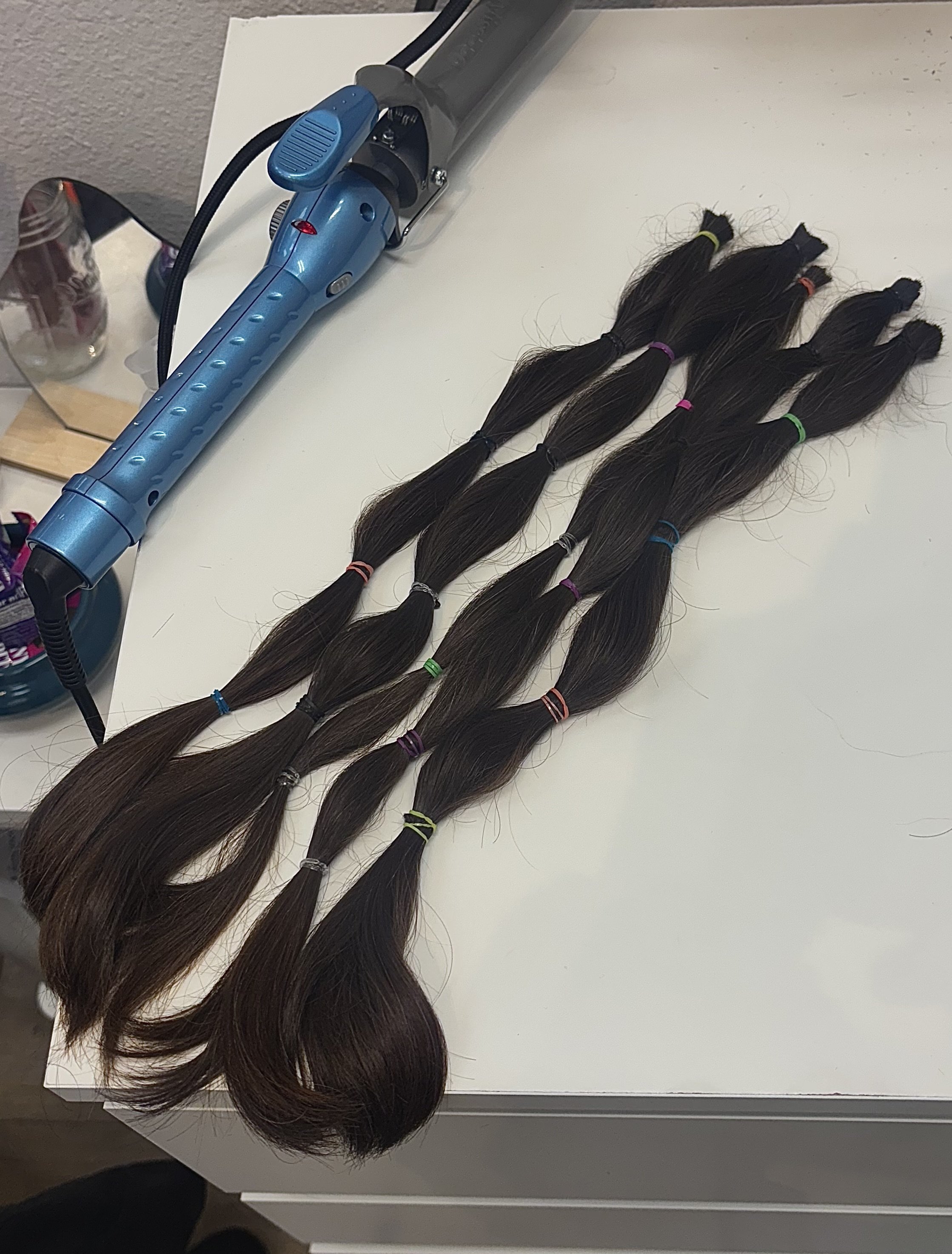 Six bundles of brown hair extensions tied with colorful bands laid out on a white table, with a blue curling iron plugged in nearby.