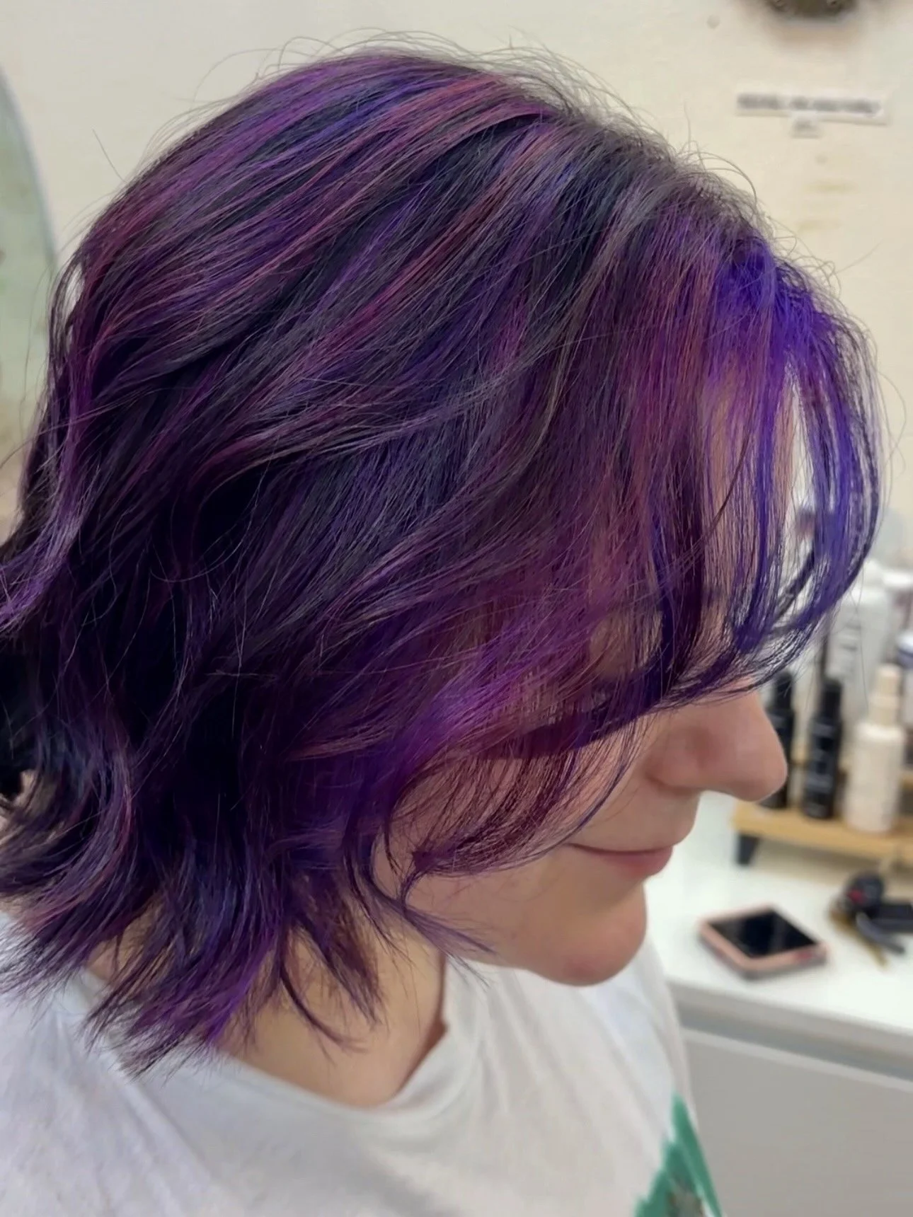 Person with straight, shoulder-length hair dyed in shades of purple and blue, standing indoors near green foliage.