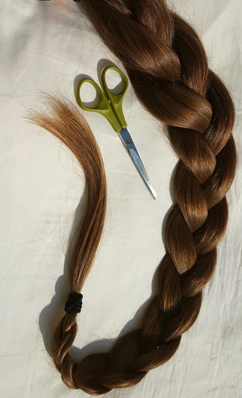 A bundle of long, brown hair with an elastic band, a pair of green scissors, and a cut strand of hair on a white surface.