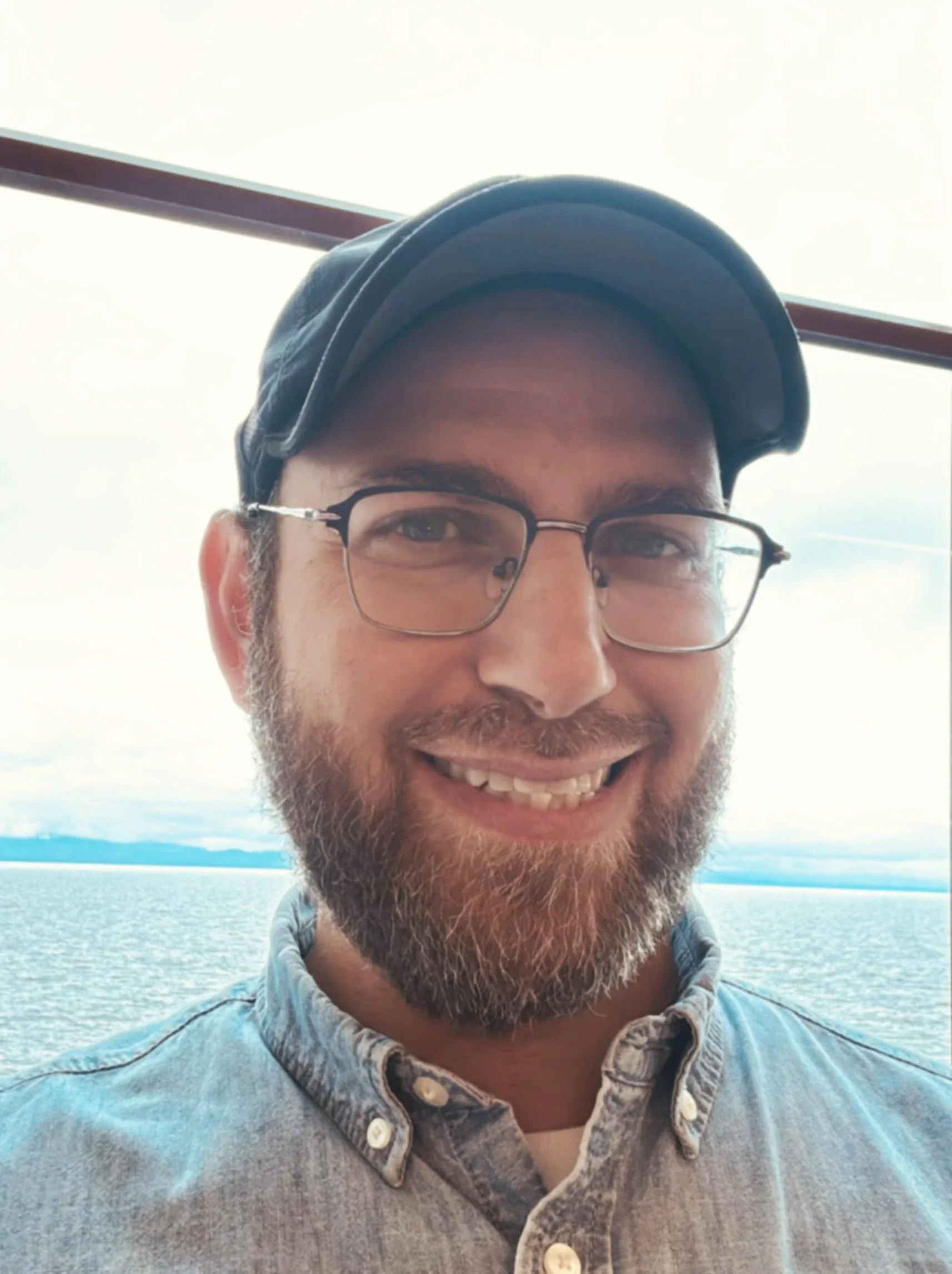 A Jewish man with glasses and a beard smiling outdoors by a large body of water, wearing a denim shirt and a cap.