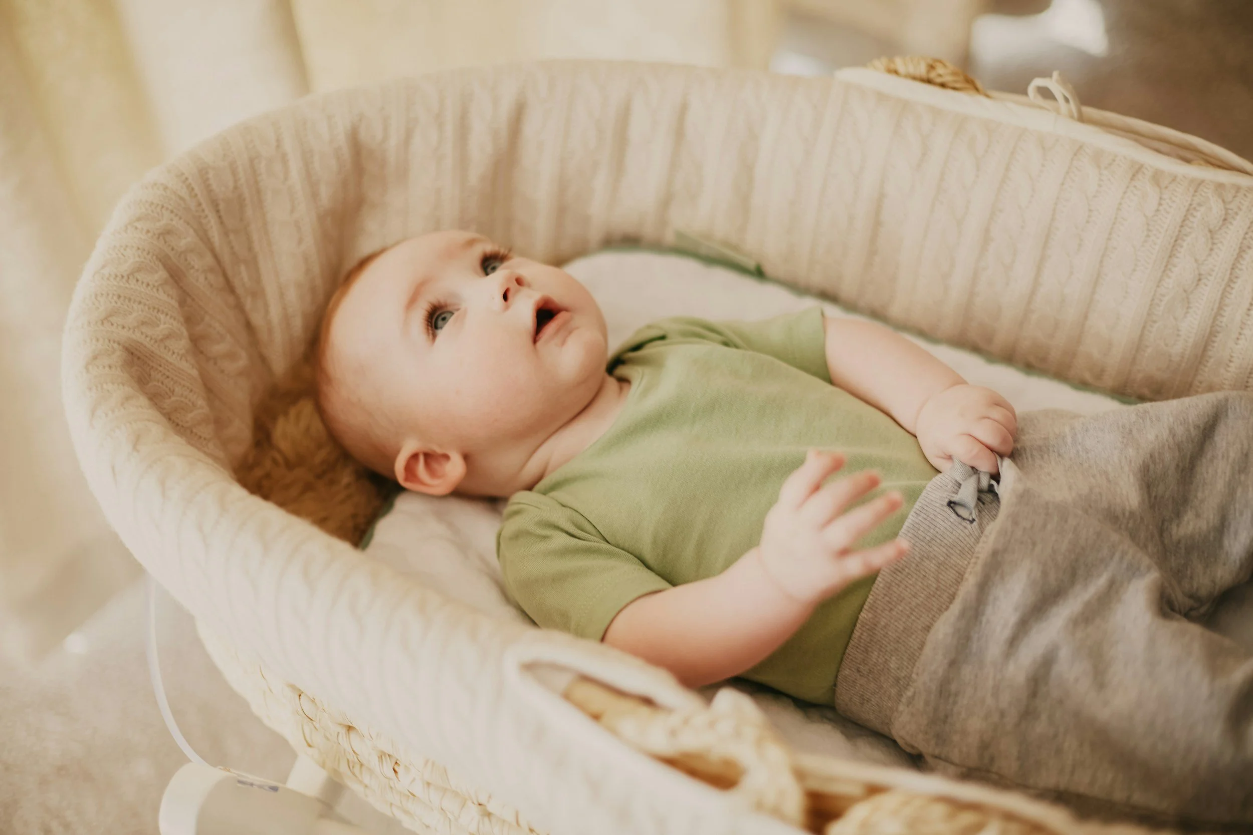 baby met groen shirtje ligt in rieten wiegje en kijkt naar boven