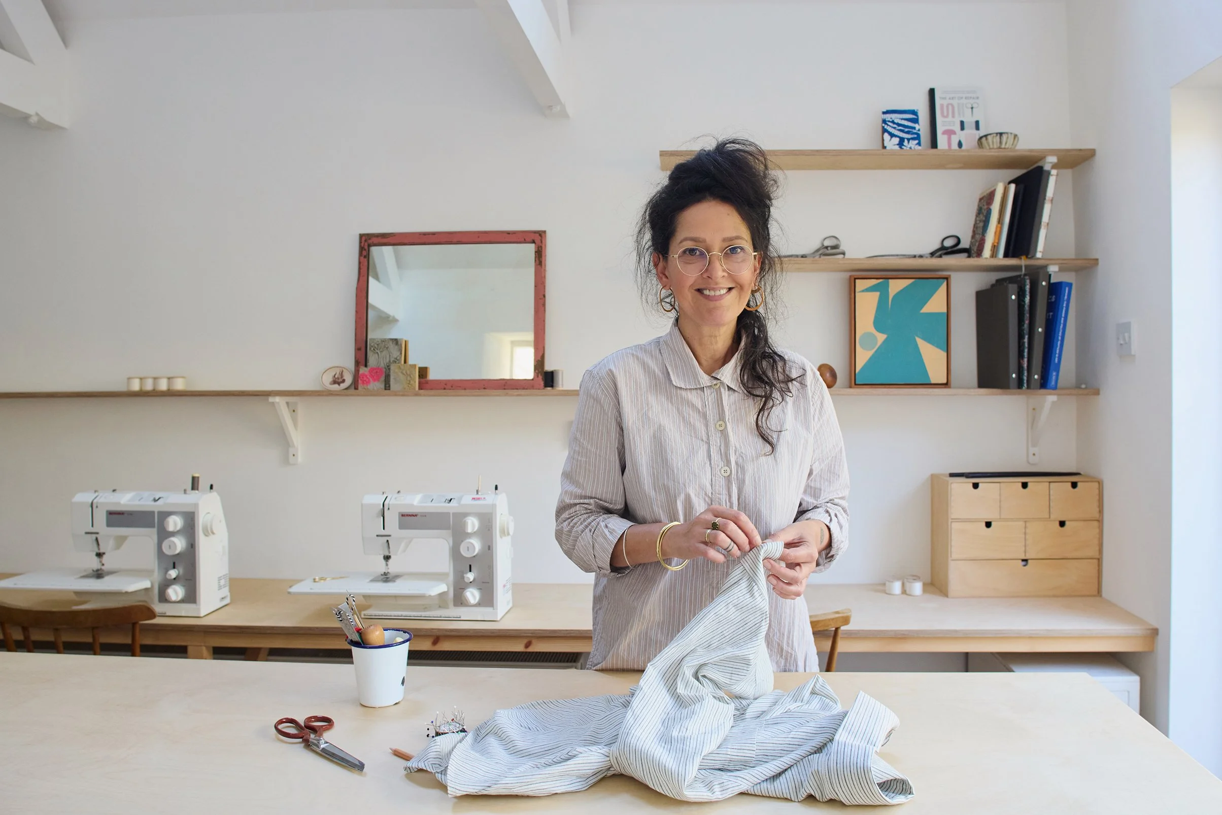 Smiling woman with glasses and earrings, standing at a worktable in a sewing room, holding a piece of striped fabric. The room has white walls, wooden shelves with books and art, and sewing machines on the countertop.