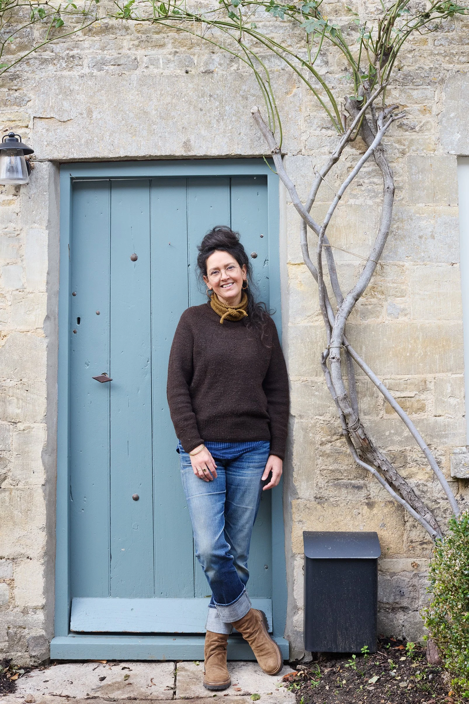 A woman with dark hair tied up, wearing glasses, a dark brown sweater, cuffed jeans, tan boots, and a yellow scarf, standing in front of a blue door with a stone wall, a small tree, and a black mailbox.