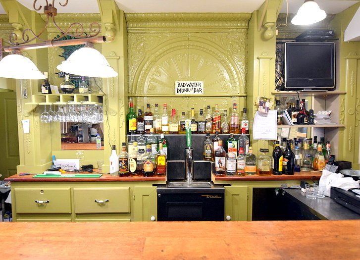 Bar area with green cabinetry, shelves with liquor bottles, glassware, a small TV, and a sign reading 'Bad Water Drink at Bar'.