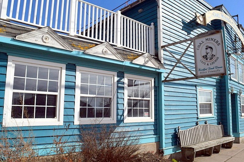 Blue building with white trim, featuring three large windows, a decorative porch trim, and a sign for 'Coal Miners Cafe & Lounge' with a vintage miner illustration, a bench in front, and part of a balcony with a blue railing above.