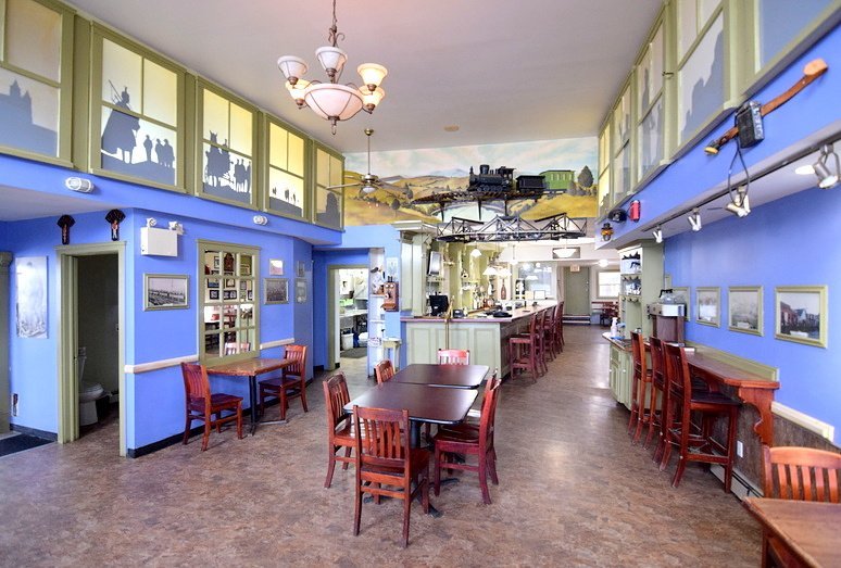 Interior of a restaurant with blue walls, wooden tables and chairs, a mural of a train and mountain landscape on the ceiling, and wall decor including framed pictures and a musical instrument.