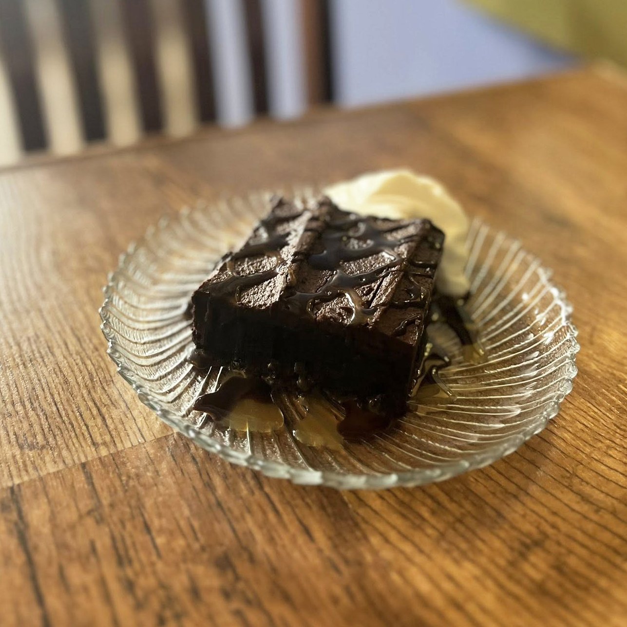 A piece of chocolate brownie with a drizzle of chocolate sauce, served with a dollop of whipped cream on a clear glass plate on a wooden table.