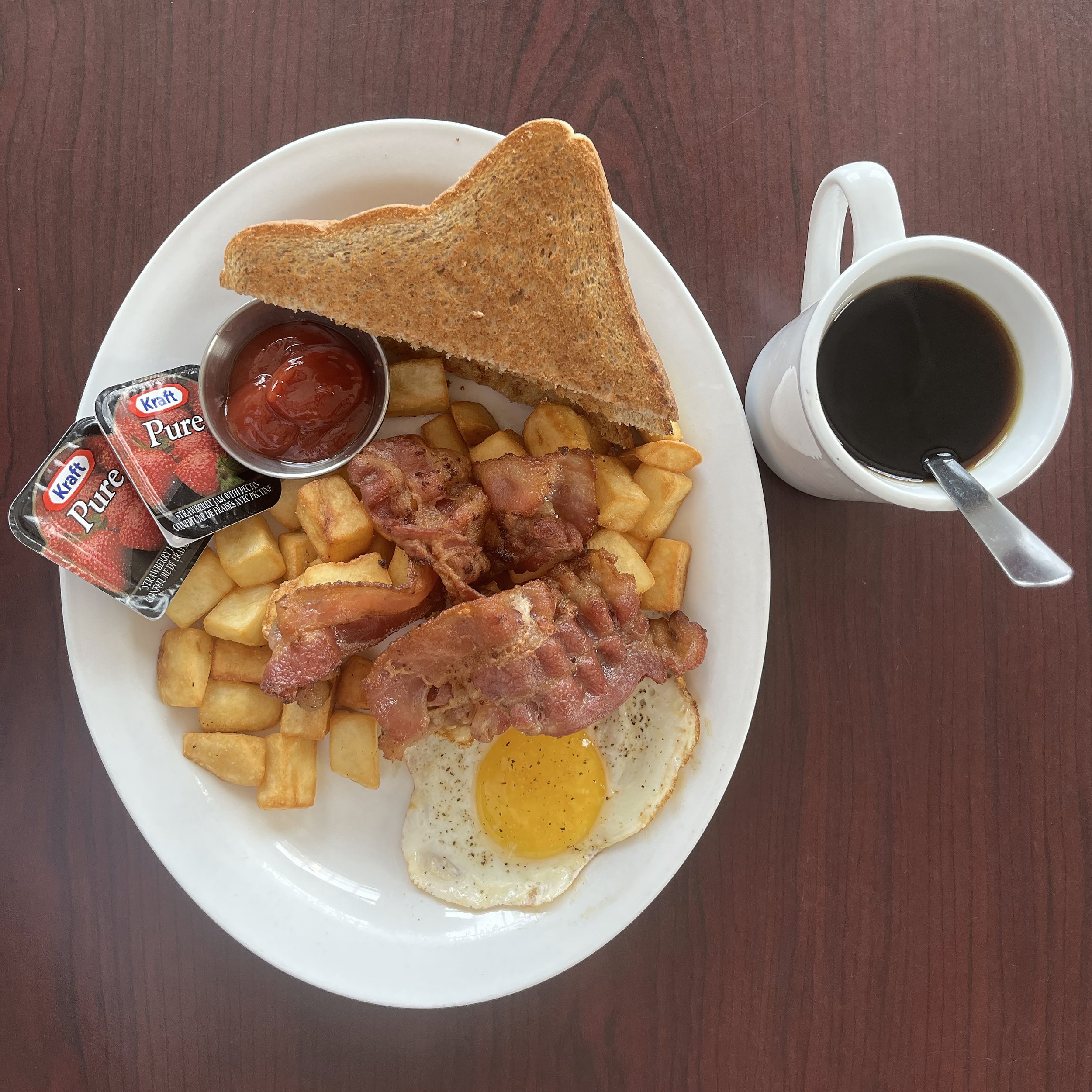 Plate with fried egg, bacon, hash browns, toast, chicken drumstick, and bacon strips, with a side of ketchup and strawberry jam, and a cup of black coffee.