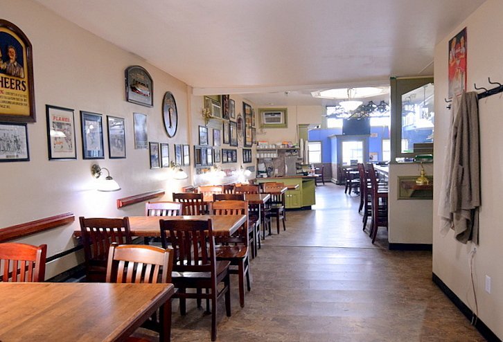 Empty restaurant with wooden tables and chairs, decorated with framed pictures and wall clocks, with a view towards the kitchen area and windows at the back.