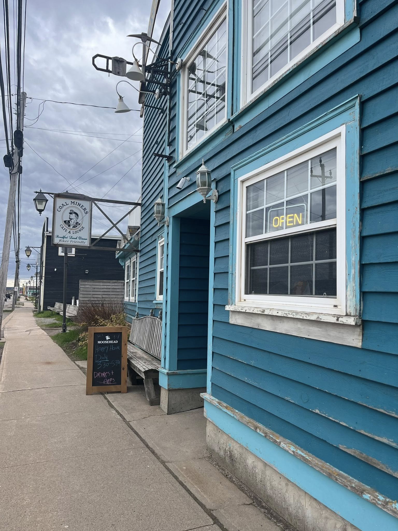 Blue wooden building with a neon 'OPEN' sign in the window, sidewalk outside, chalkboard sign, and a sign for Coal Miners Cafe & Lounge in the background.
