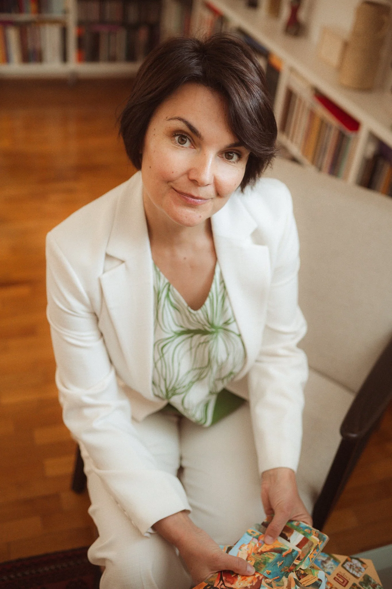 A woman with short dark hair sitting indoors, holding and looking at a stack of colorful photo cards or postcards. She is dressed in a white blazer and a green and white striped top, with bookshelves filled with books in the background.
