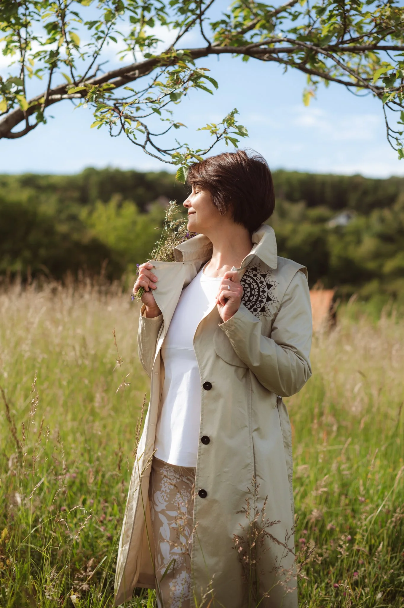 A woman with short brown hair stands in a field of tall grass and wildflowers, smiling with her eyes closed, holding a small bouquet of flowers, with green trees in the background and blue sky above.