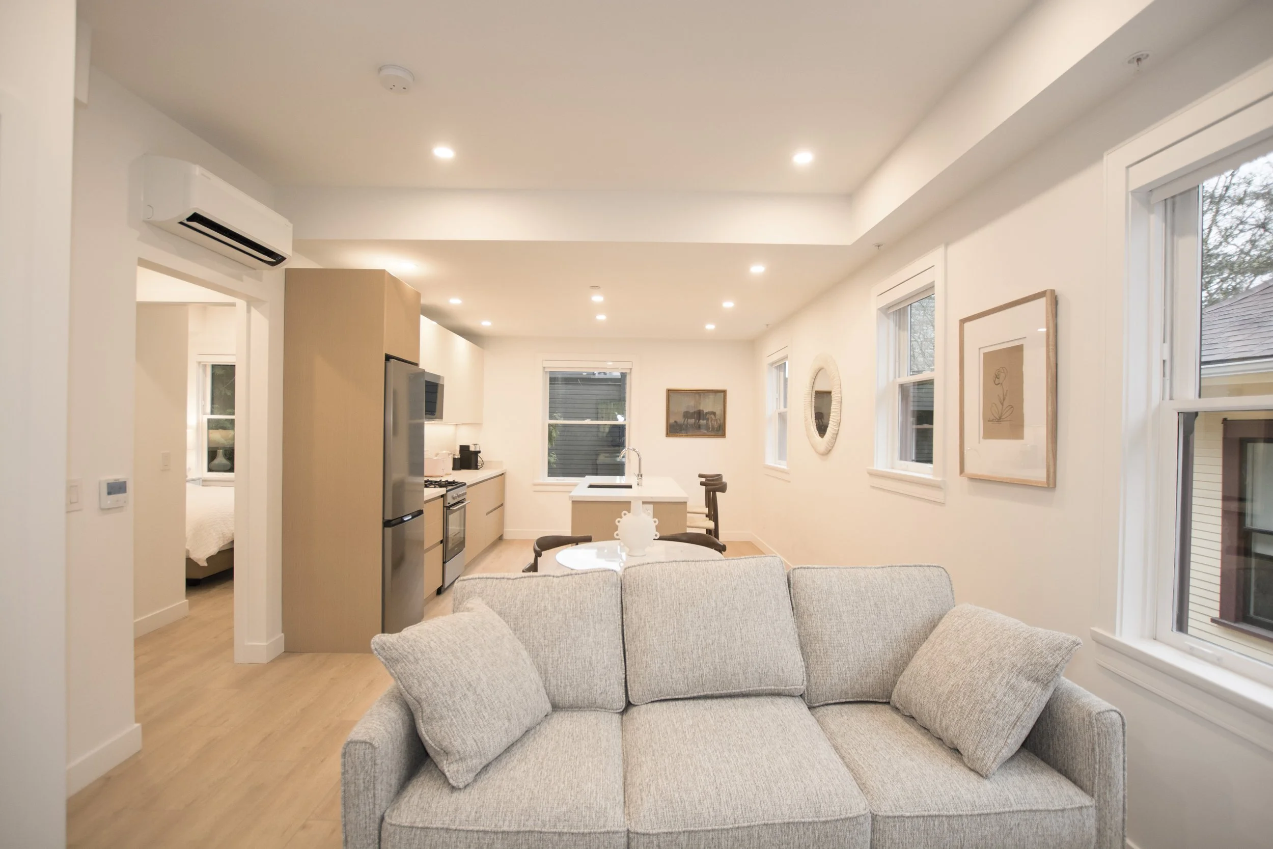 Living room with beige sofa, light wood flooring, white walls, and picture frames. Open kitchen with refrigerator, stove, and sink. Multiple windows with view outside, artwork on walls, and ceiling lights.