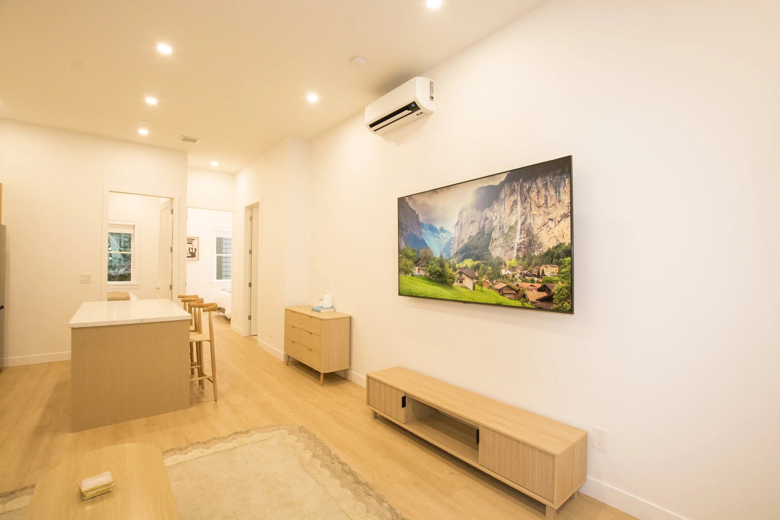 Modern living room with light-colored walls, wooden furniture, a wall-mounted TV displaying a mountain landscape, air conditioning unit, and a bright, minimalist decor.