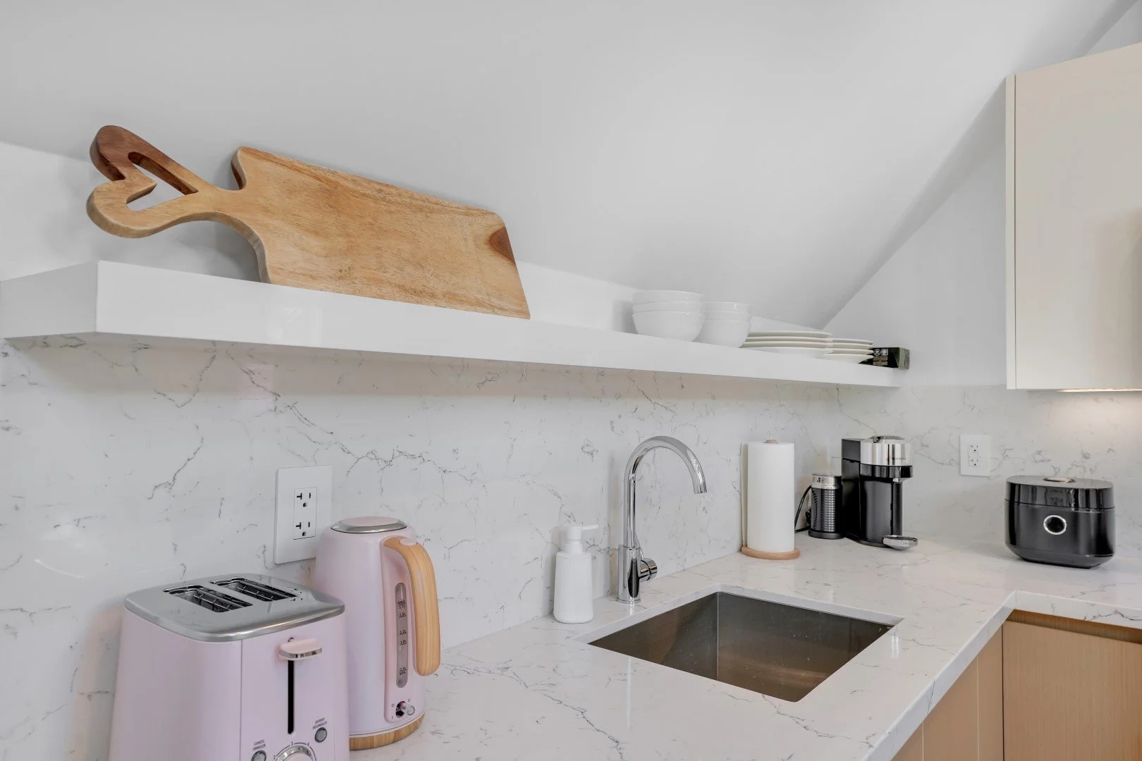 Kitchen countertop with a pink toaster and kettle, black coffee maker, paper towel holder, white bowls and plates on a white shelf, and wooden cutting board on the shelf.