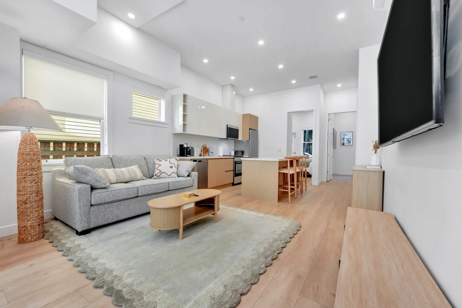 Open-concept living room and kitchen with light wood flooring, gray sofa, beige rug, flat-screen TV, and minimalist decor.
