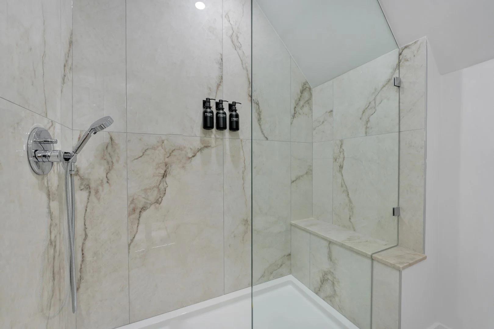 Interior of a modern shower with marble tiles, a handheld showerhead, and three black dispensers on the wall, with a built-in marble bench.