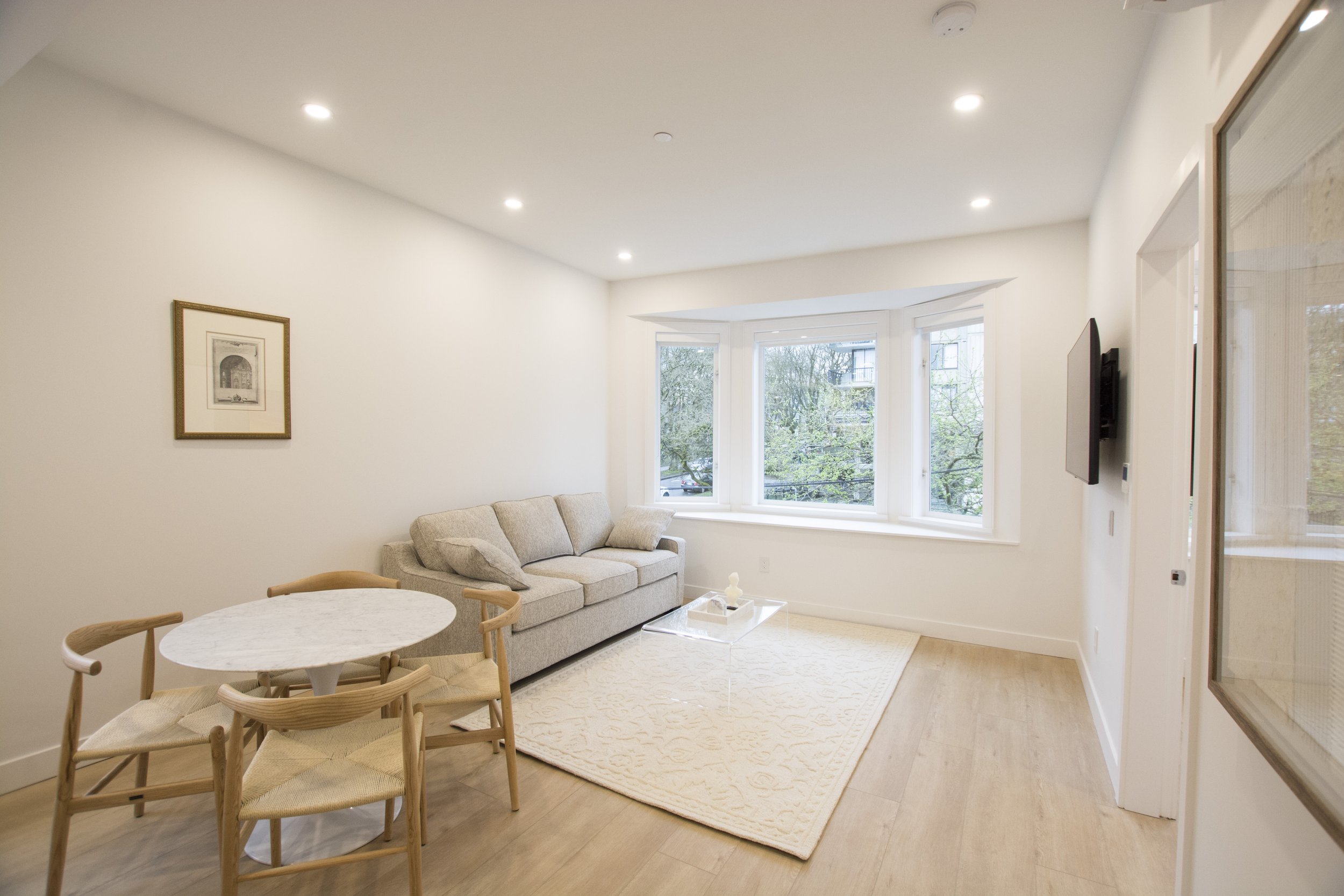 Bright living room with light-colored sofa, round white marble dining table with four wooden chairs, large bay window showing trees outside, wall-mounted TV, framed artwork, beige rug on light wood flooring, and white walls.