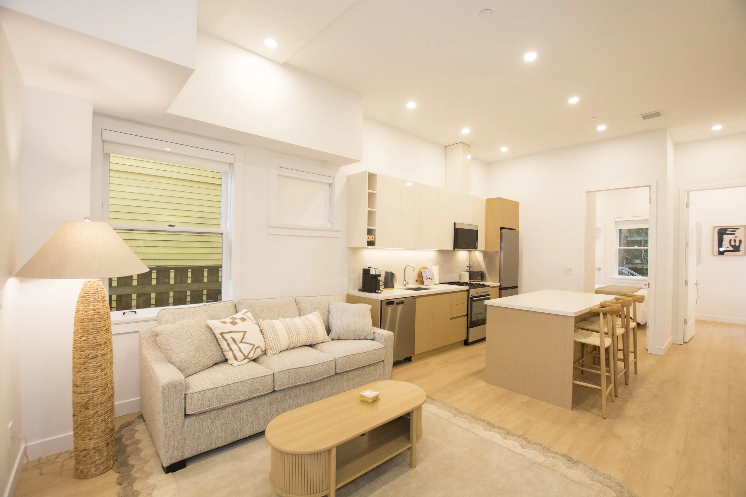 Open-concept living room and kitchen in a modern apartment with beige sofa, wooden coffee table, kitchen island with barstools, and white cabinets, illuminated by recessed ceiling lights.