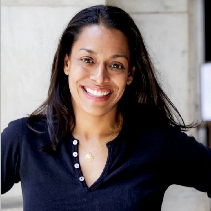 Smiling woman with dark hair wearing a navy blue top and gold necklace, standing outdoors with a neutral background.