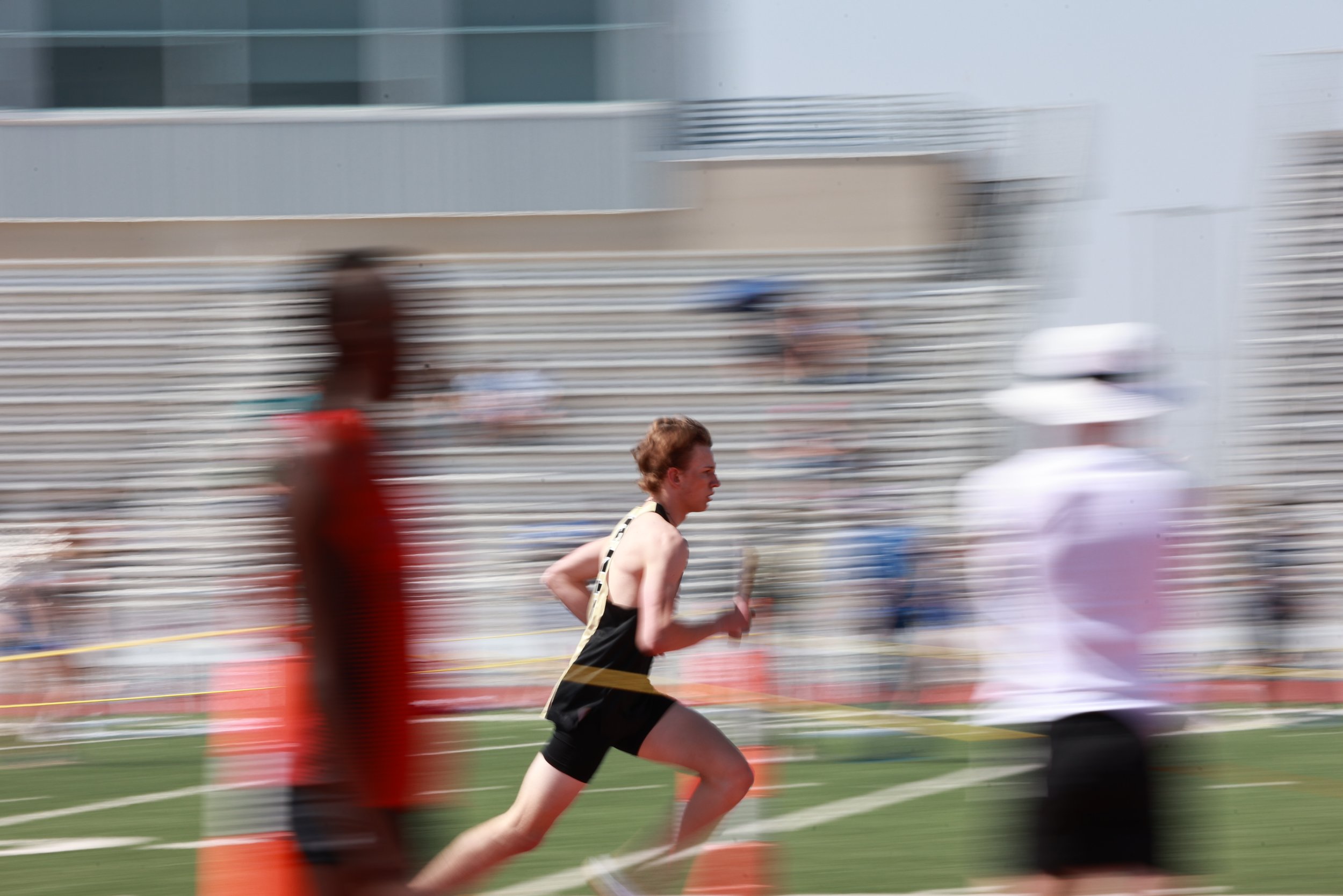 A male athlete running in a track event with spectators in the background during daytime, motion blur indicating speed.