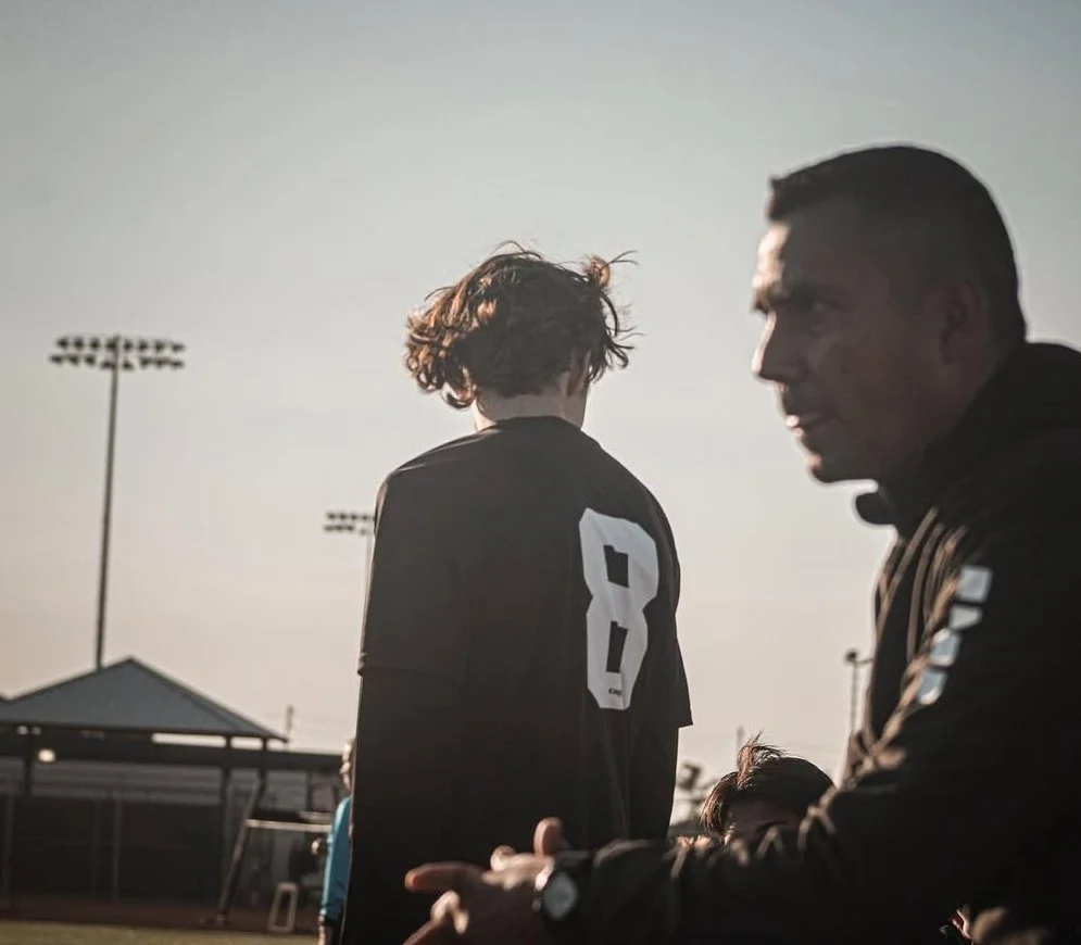 A young football player in a black jersey with the number 8 on the back, standing on the field, with an adult coach or teammate nearby, during a game at sunset.
