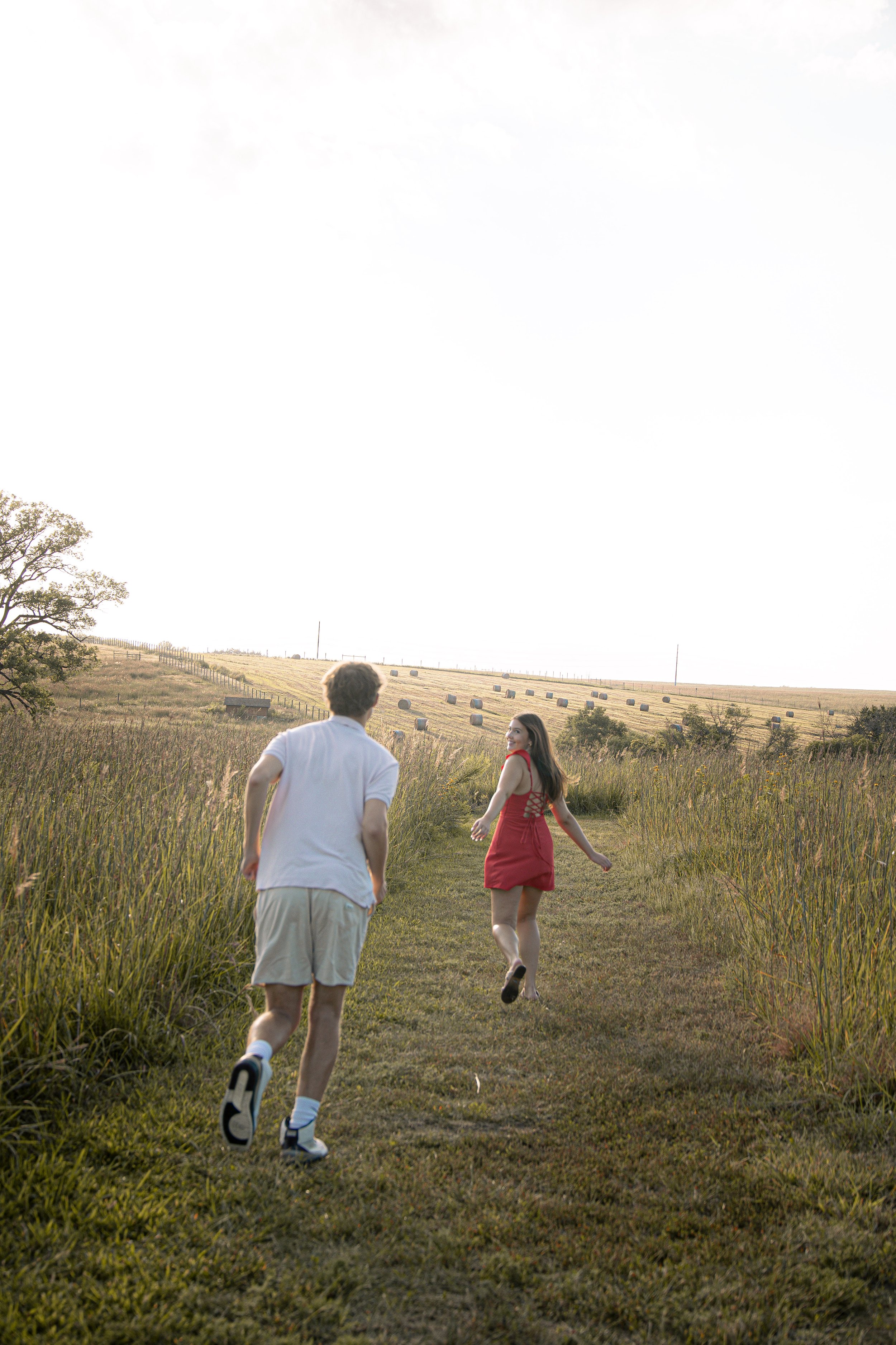 A young man and woman running on a grassy trail through open fields with hay bales in the distance during daytime.