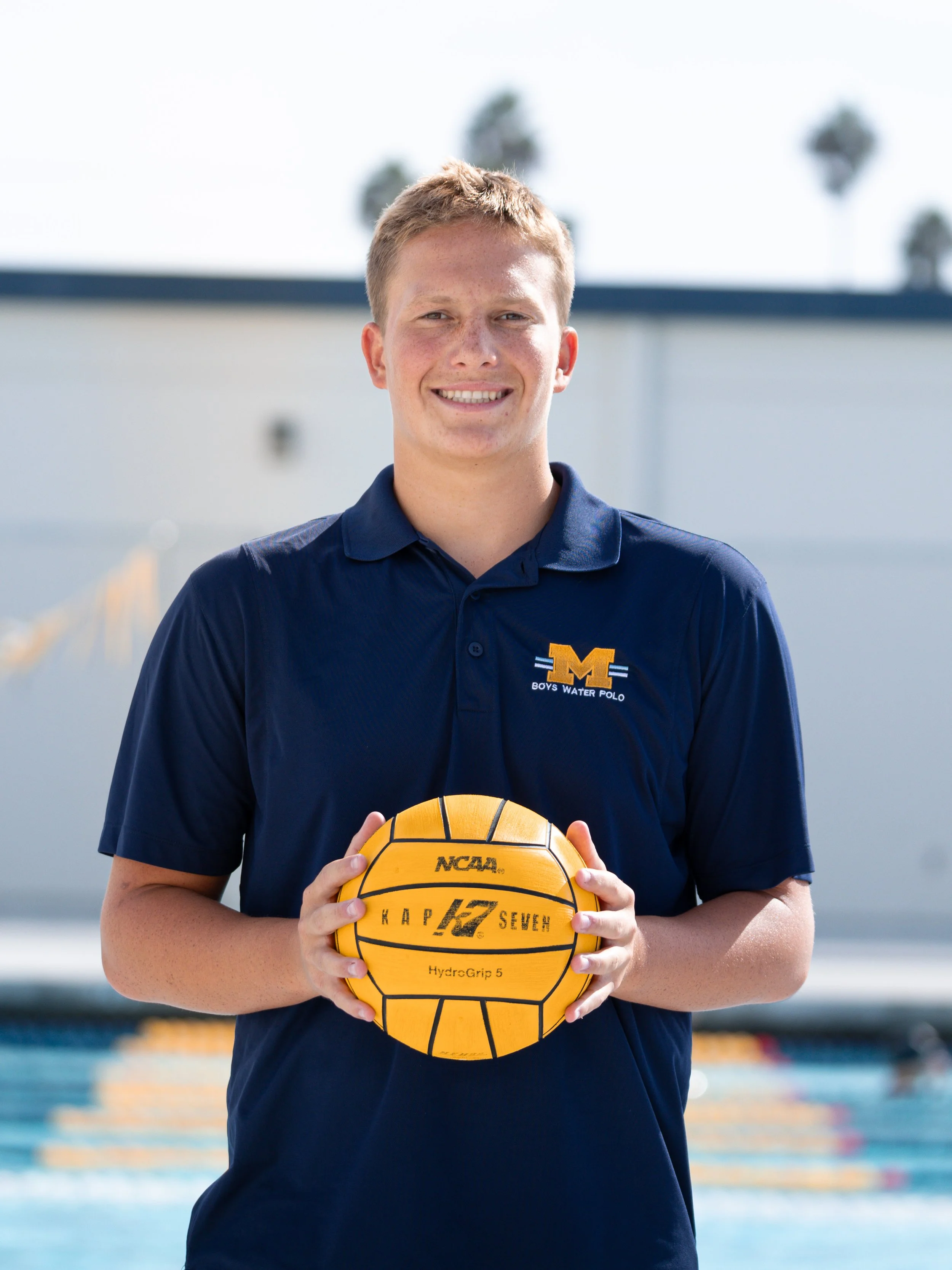 Young male water polo player in navy polo shirt holding a yellow water polo ball in a pool area.