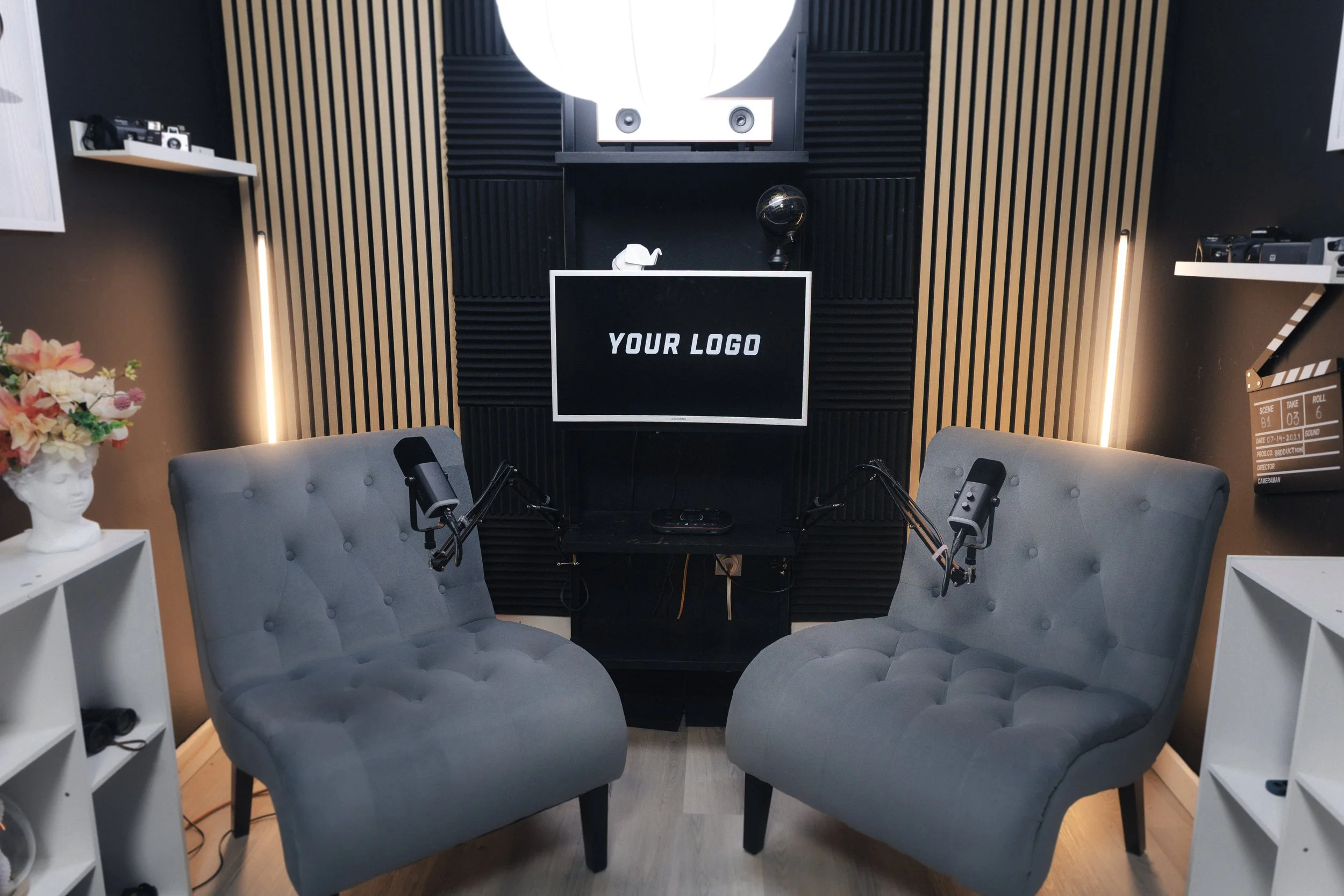 Podcast studio with two gray tufted chairs, a digital monitor with 'Your Logo', microphones, soundproofing panels, shelves with equipment, and decorative items.